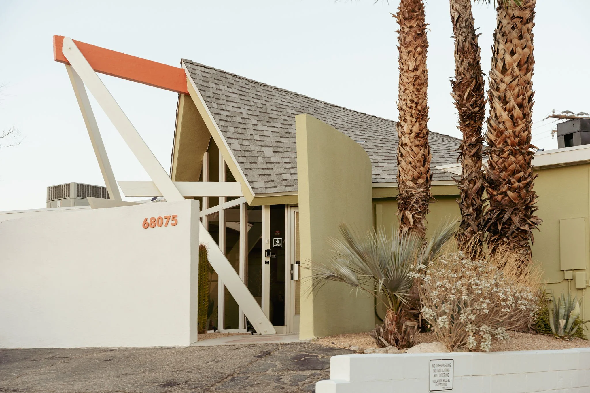 A modern building with a gray shingled roof, yellow walls, and a white angled support structure. There are three tall palm trees to the right of the building and desert plants in the foreground. A sign on the white barrier says 'No Trespassing, No Solicitations, No Loitering.'