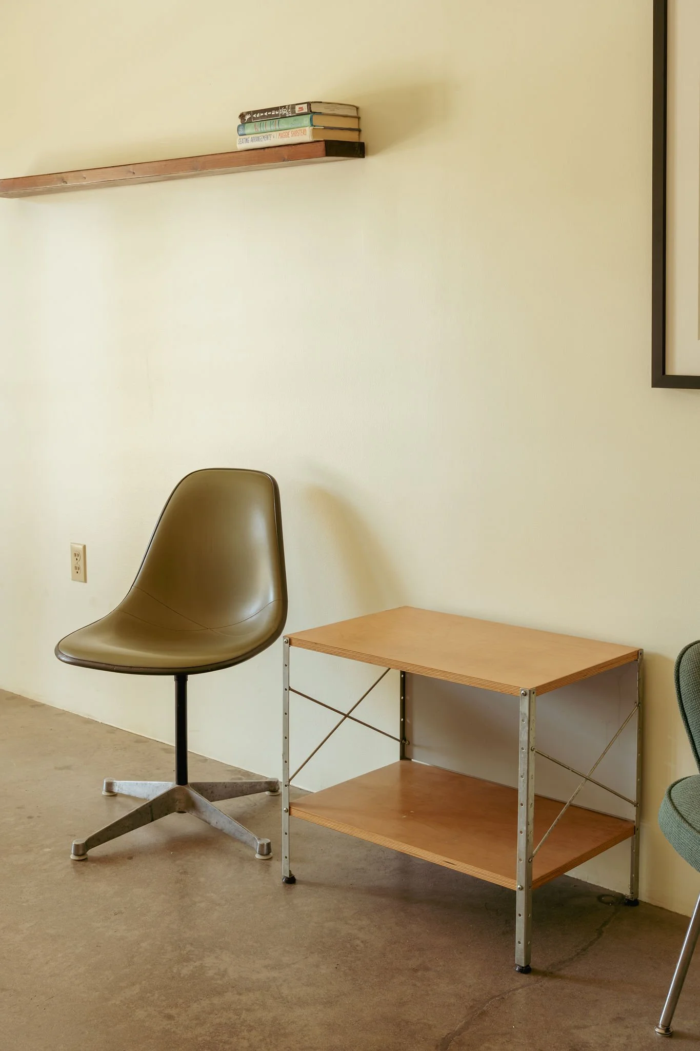 A beige office chair with a rounded back and metal base, positioned in front of a wooden and metal shelving unit with three shelves, next to a small wooden table in a room with off-white walls and a concrete floor.