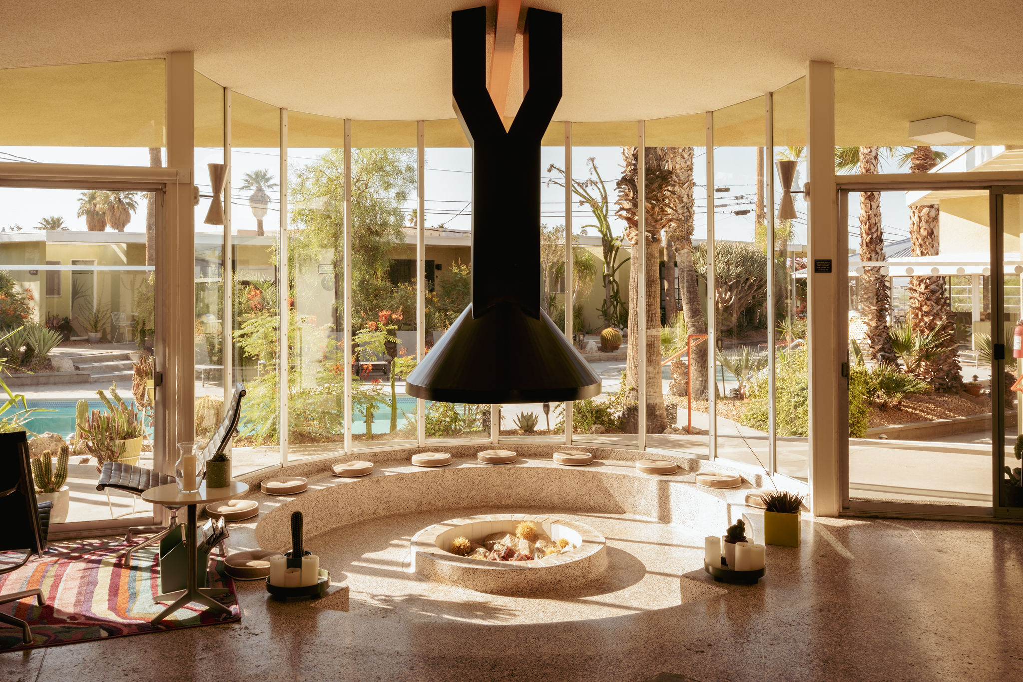 Interior view of a modern living space with large floor-to-ceiling windows, a black fireplace in the center, a circular seating area with beige cushions around a fire pit, and outdoor palm trees and plants visible through the windows