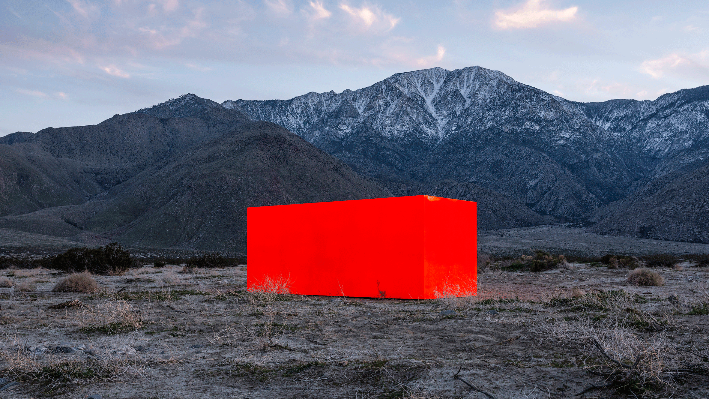 A mountain range with snow-capped peaks and a cloudy sky, with a large red cube-shaped object in the foreground on a desert landscape.