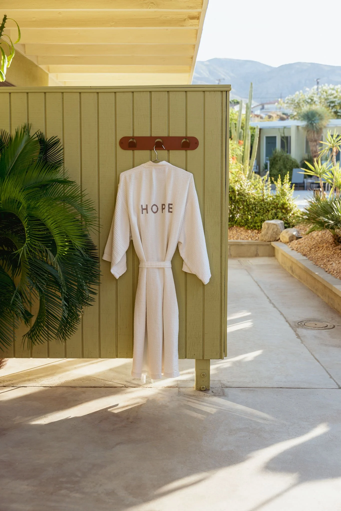 White bathrobe with the word 'HOPE' on the back hanging on a wooden fence in an outdoor patio area with plants and mountains in the background.