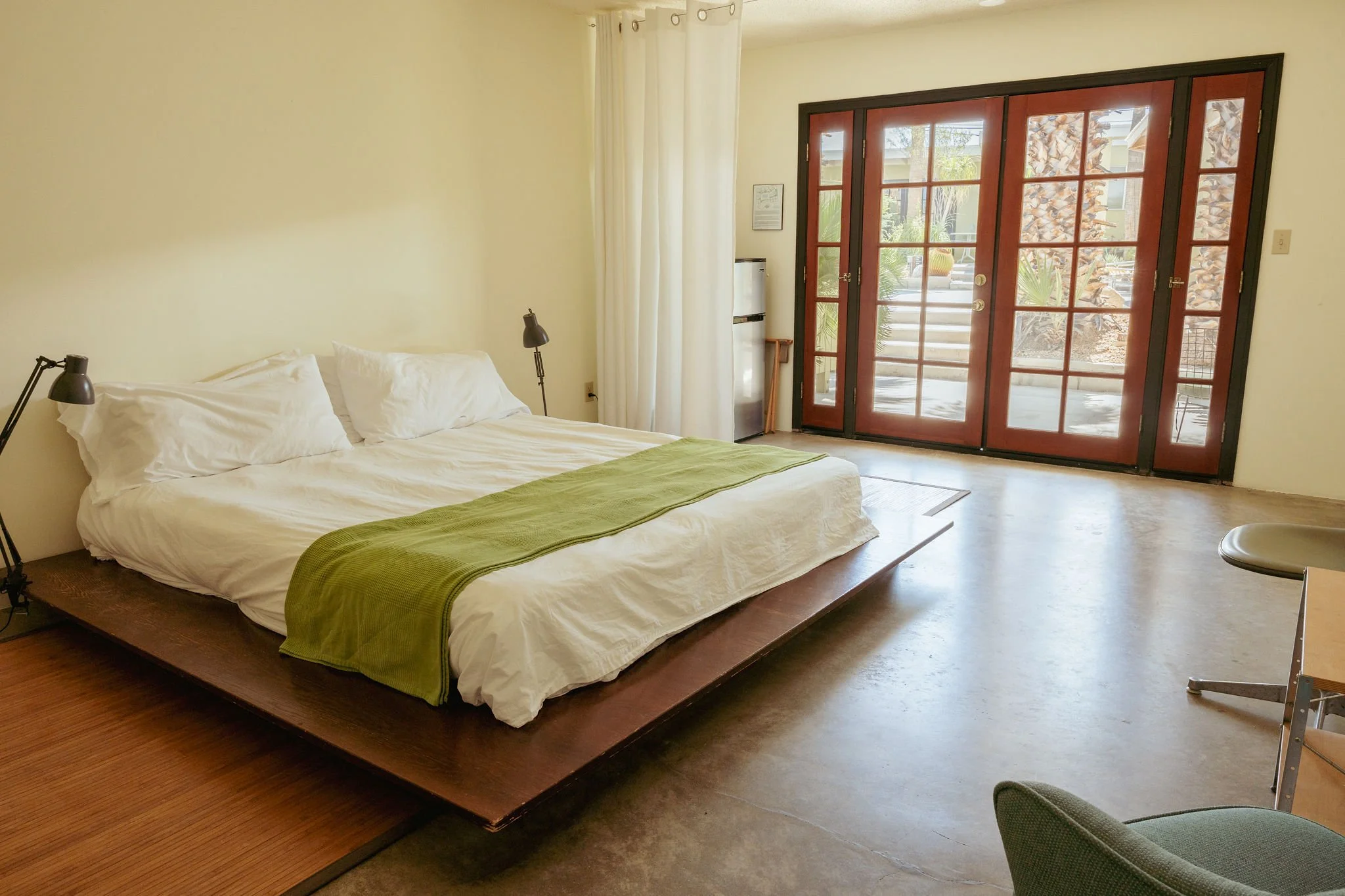 A bedroom with a wooden bed frame, white bedding, and a green throw blanket. Two black bedside lamps, cream curtains, and a large set of glass-paned wooden doors leading outside. Outside, desert plants and a staircase are visible.