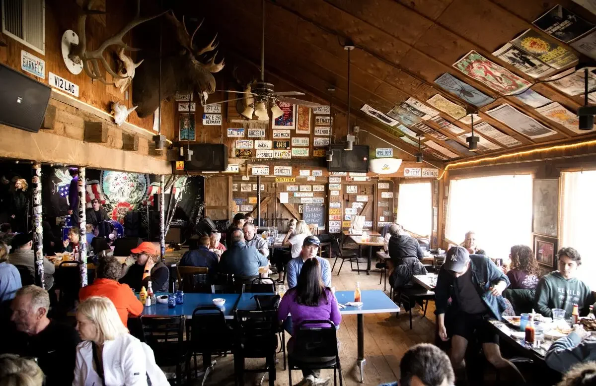 Interior of a busy restaurant or bar with wooden walls and ceiling, decorated with mounted animal heads, license plates, and artwork. Several groups of people are sitting at tables, dining and socializing. The space has large windows letting in natural light.