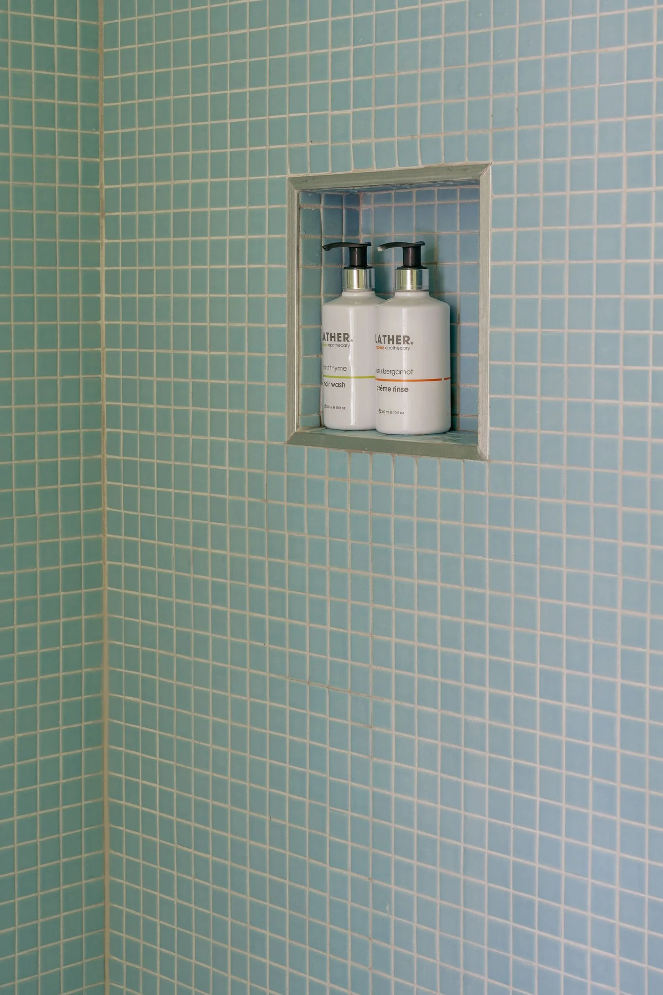 Two bottles of bathroom toiletry products placed in a recessed shelf within a tiled shower wall. The tiles are small, light blue, with white grout.