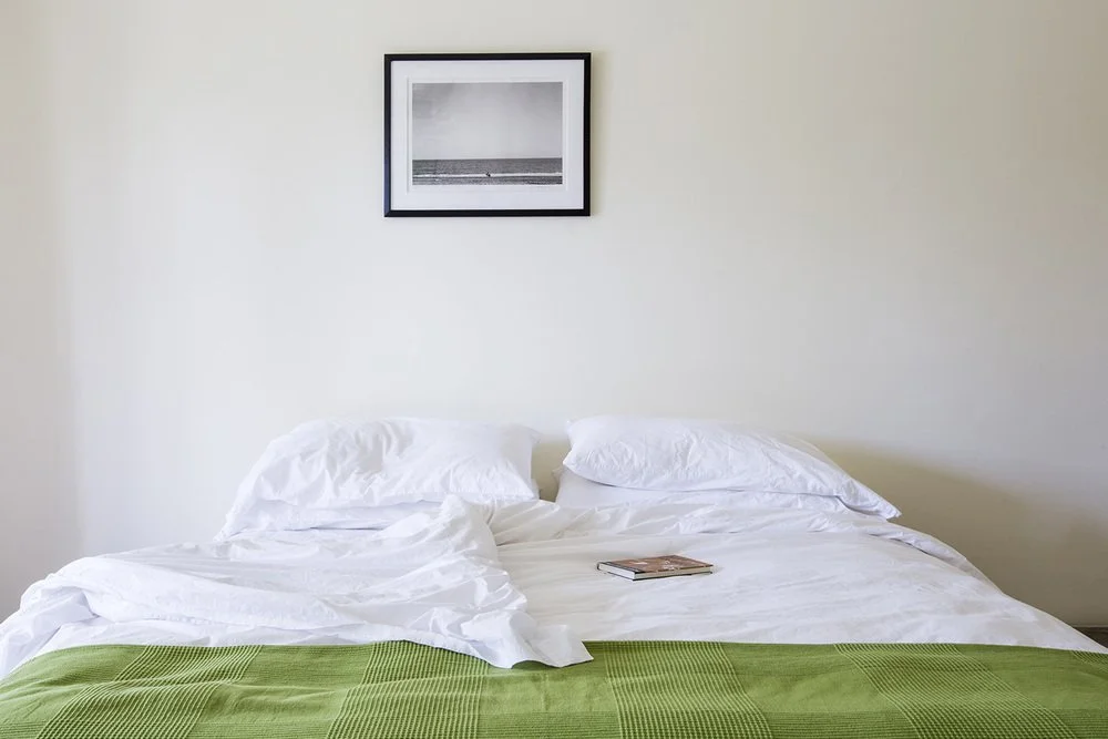 A neatly made bed with white sheets, pillows, and a green blanket at the foot, positioned against a beige wall with a framed black-and-white beach photograph hanging above.