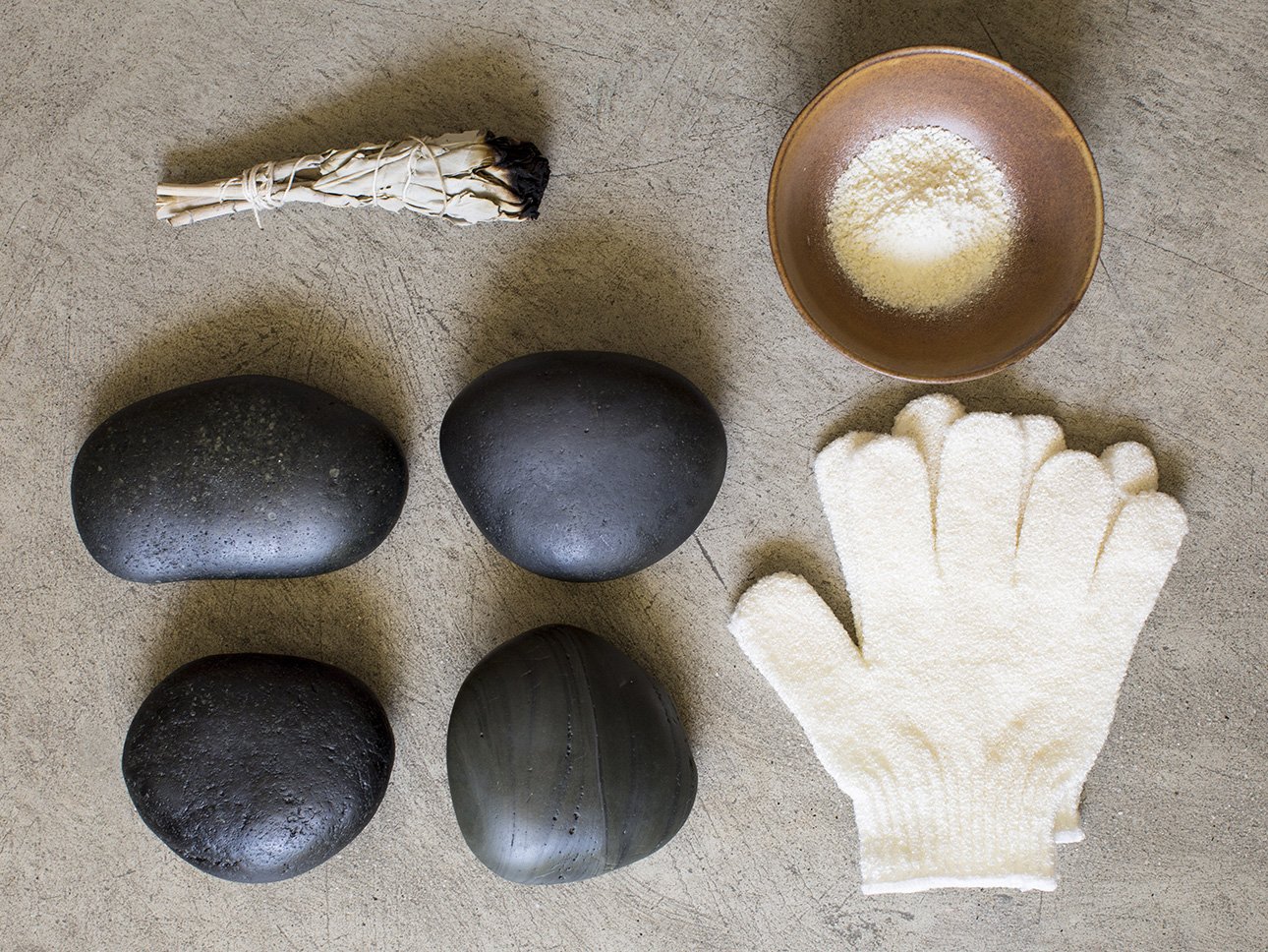 A collection of five black stones, a rolled bundle of sage, a small bowl of white powder, a pair of white textured gloves, and a metallic ball.