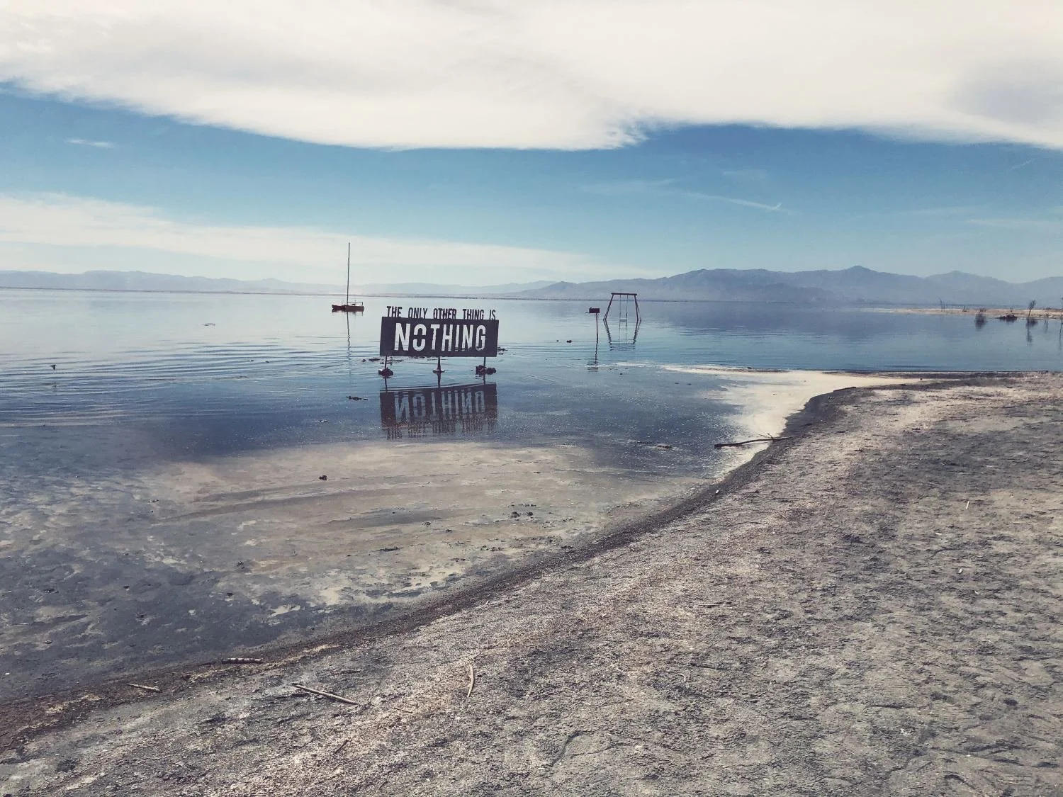 A sign in shallow water near a sandy shoreline reads, 'The only other thing is nothing.' In the background, there are boats and a swing set, with mountain ranges on the horizon under a cloudy sky.