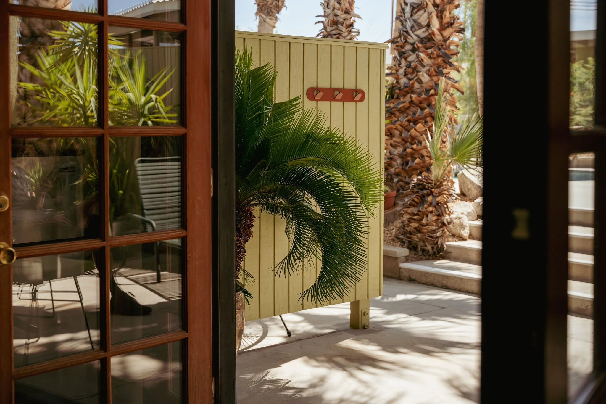 View through a glass door showing an outdoor patio area with palm trees, potted plants, a yellow wooden fence with hooks, and a concrete staircase leading up.