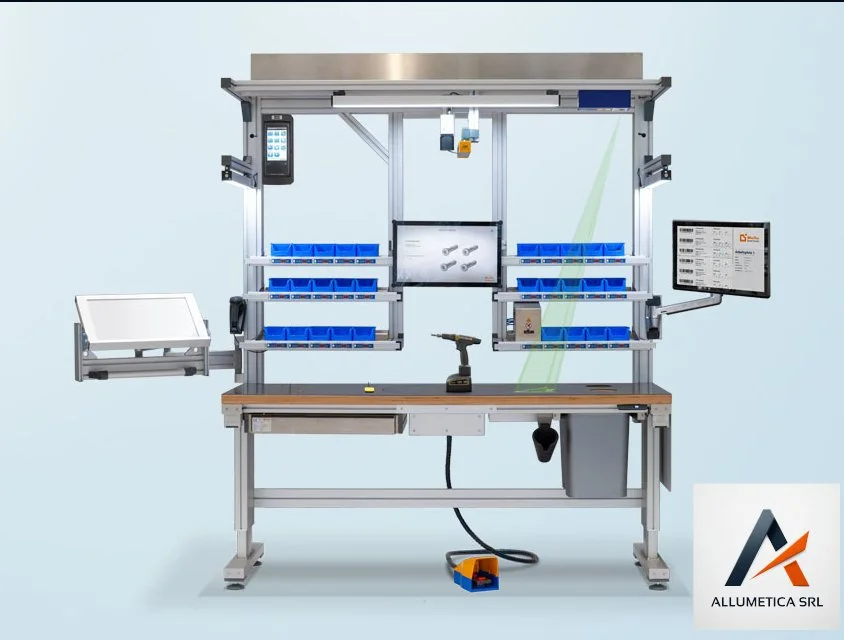 Postazione di lavoro con braccio robotico, computer, divise di stoccaggio con contenitori blu, e strumenti per assemblaggio.