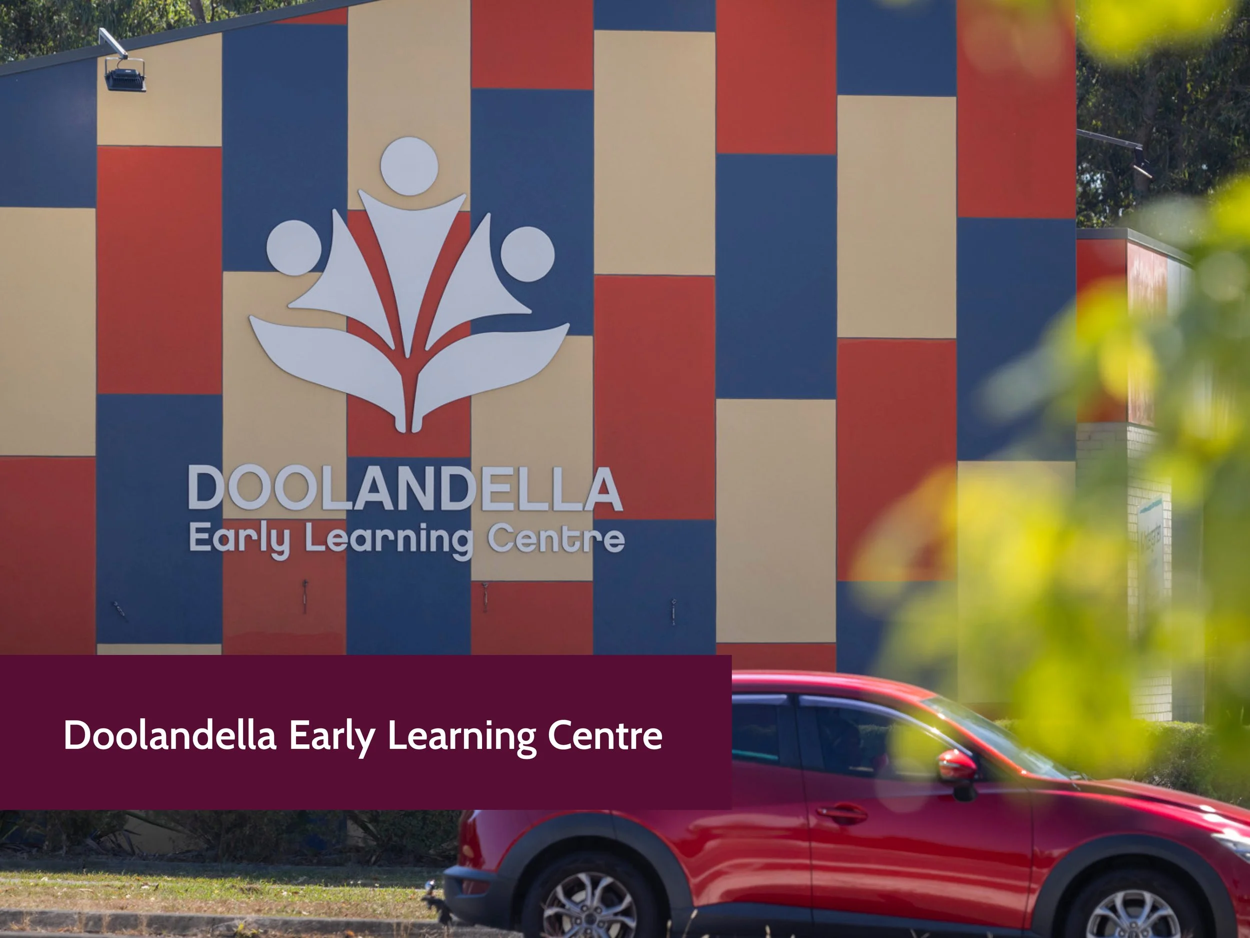 A colorful building with blue, red, and beige checkered pattern and a large logo of a white lotus flower with three circular figures, signifying Doolandella Early Learning Centre, is visible. There is a red car parked in front of the building, partia