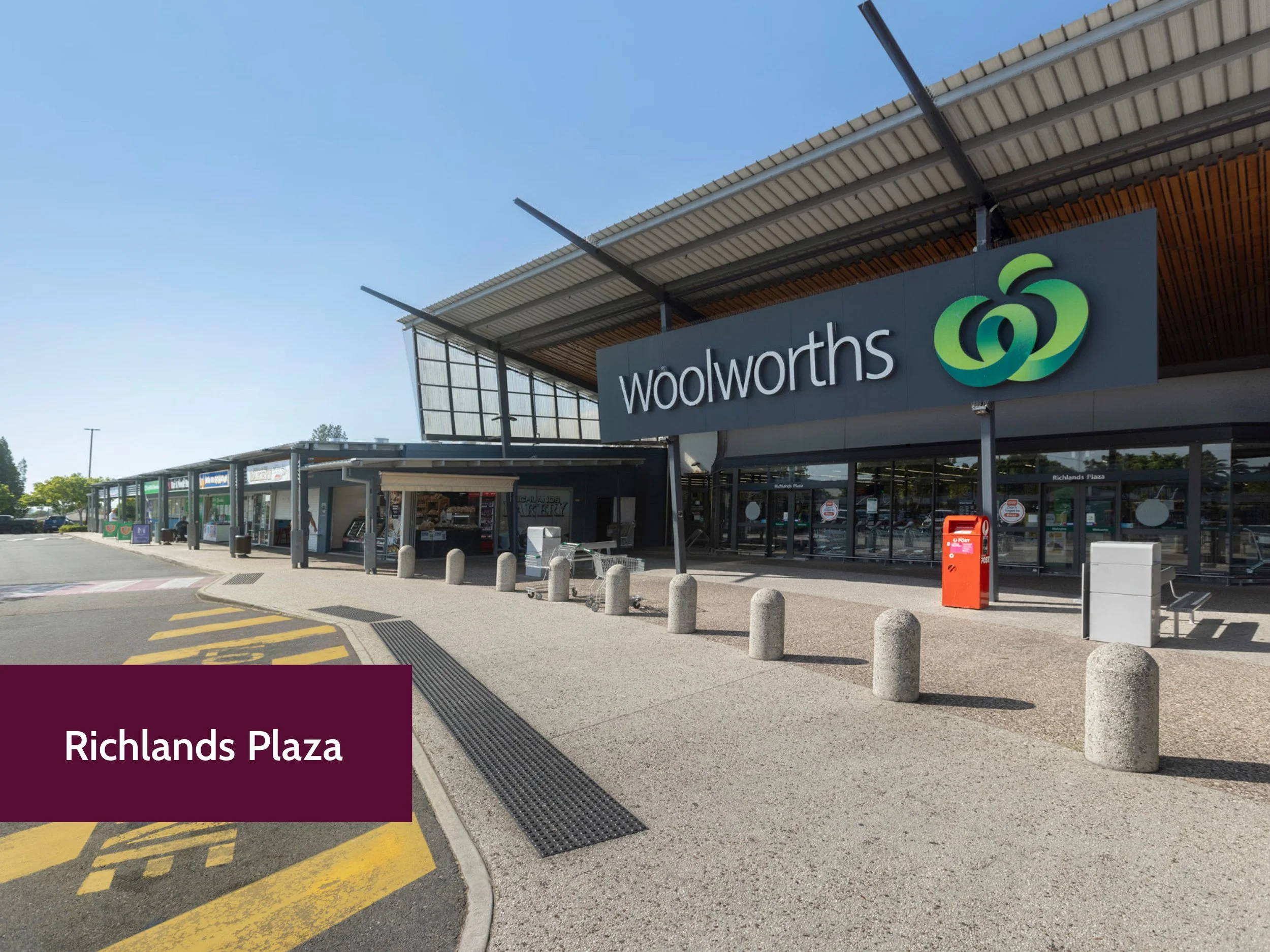 The exterior of a Woolworths store at Richlands Plaza, showing the store sign, shopping carts, and a few small shops outside the entrance on a sunny day.