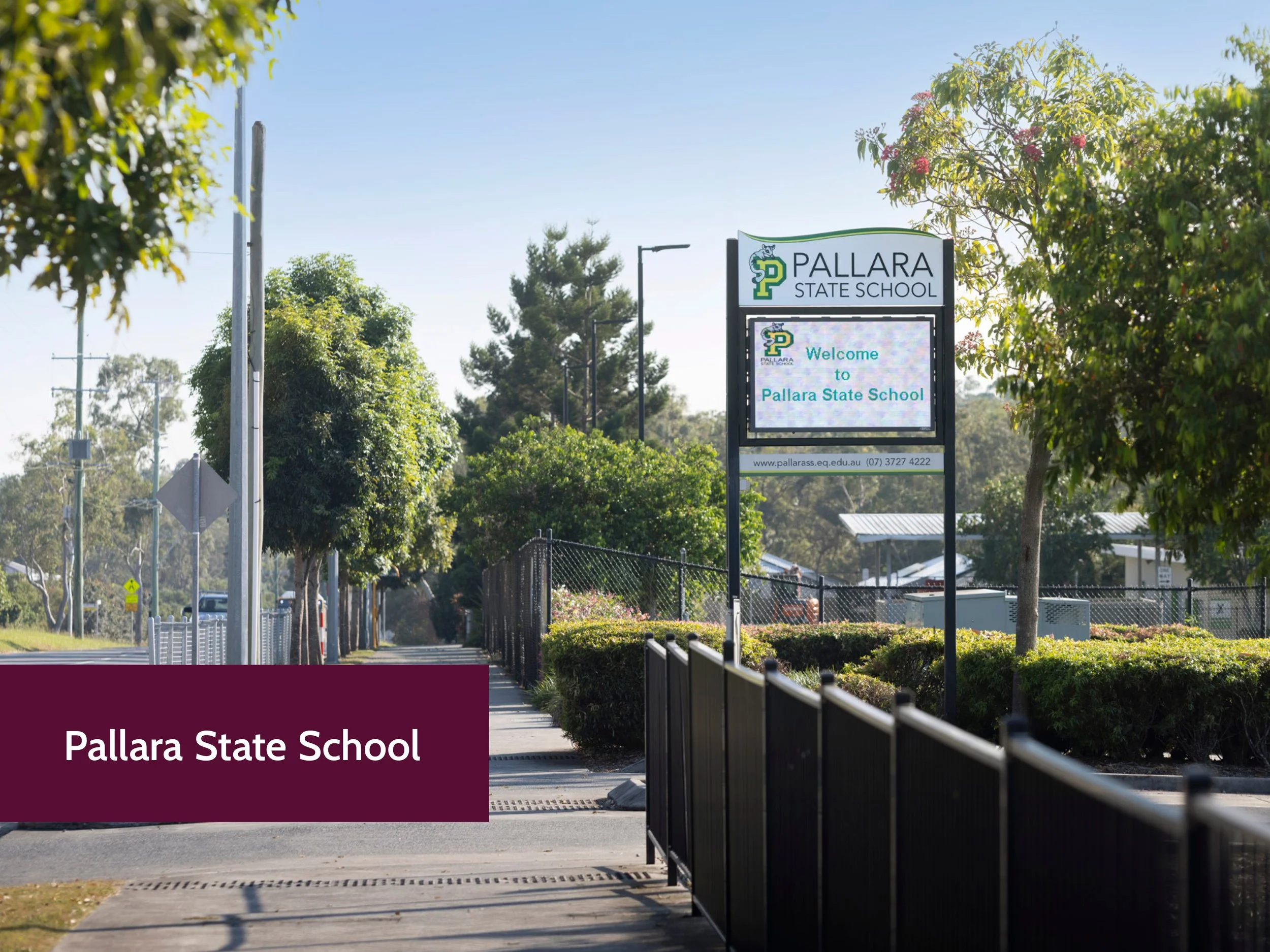 Signboard at Pallara State School surrounded by trees, with a sidewalk and fence in front, and utility poles on the left, under a clear blue sky.