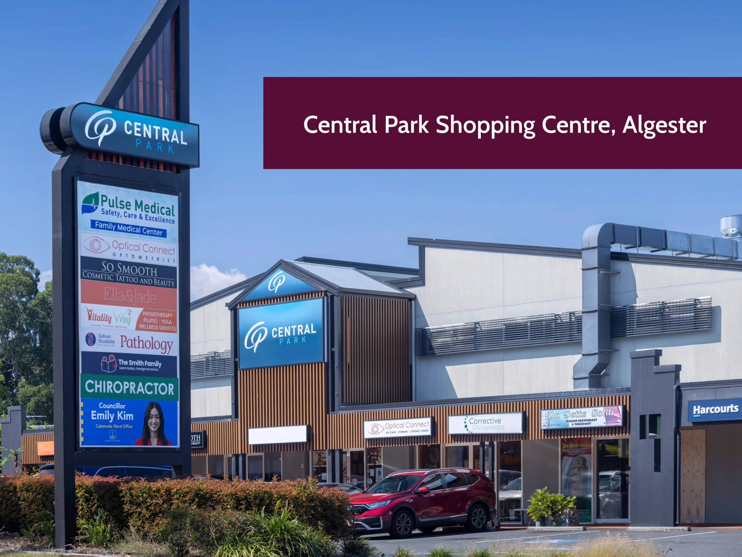 A shopping center with signs for various businesses including Pulse Medical, Optical Connect, and a chiropractic clinic. A large sign with the shopping center's name, Central Park, is visible at the top of the signpost. The building has multiple stor
