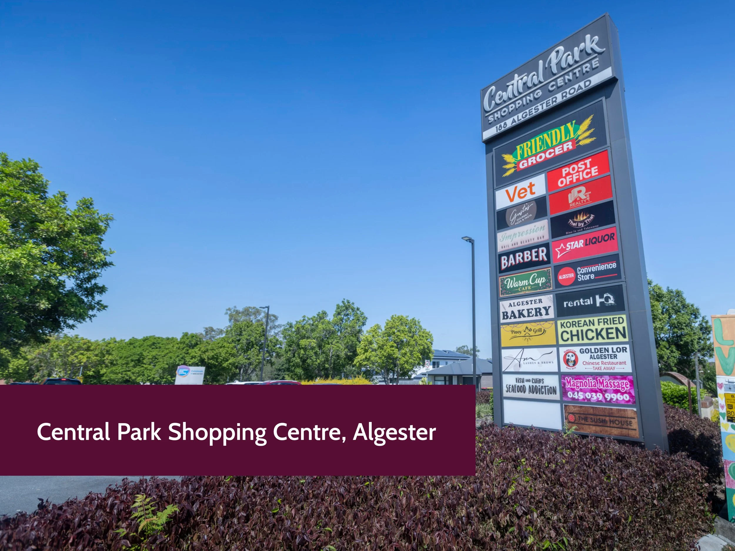 A tall signpost with various business advertisements in a parking lot, under a clear blue sky, with green trees and parked cars in the background.
