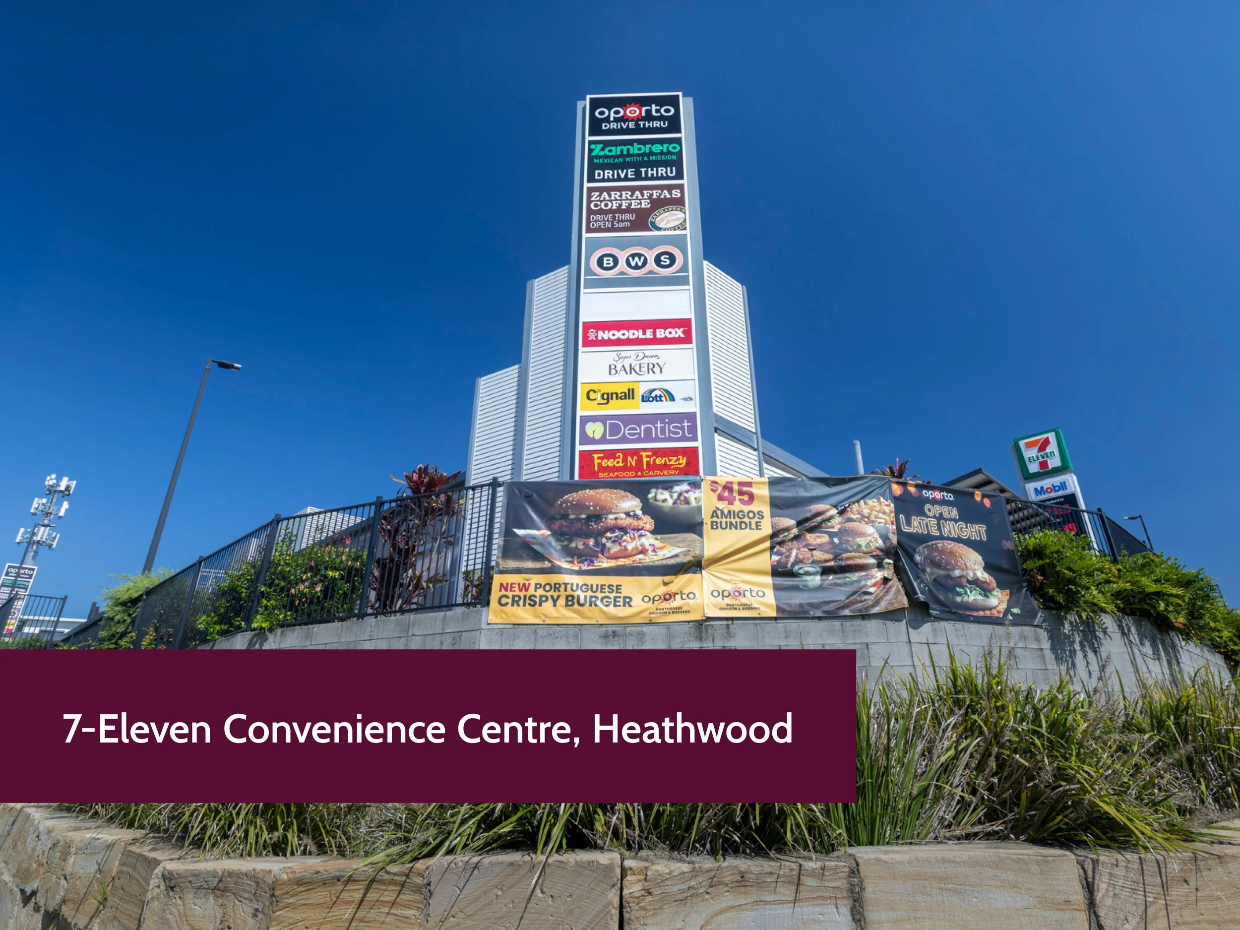 A tall sign with various store logos, including Opporto, Zambrero, Zarraffa's Coffee, BWS, Noodle Box, a bakery, Cignall, and 7-Eleven, against a clear blue sky near the 7-Eleven Convenience Centre in Heathrow.
