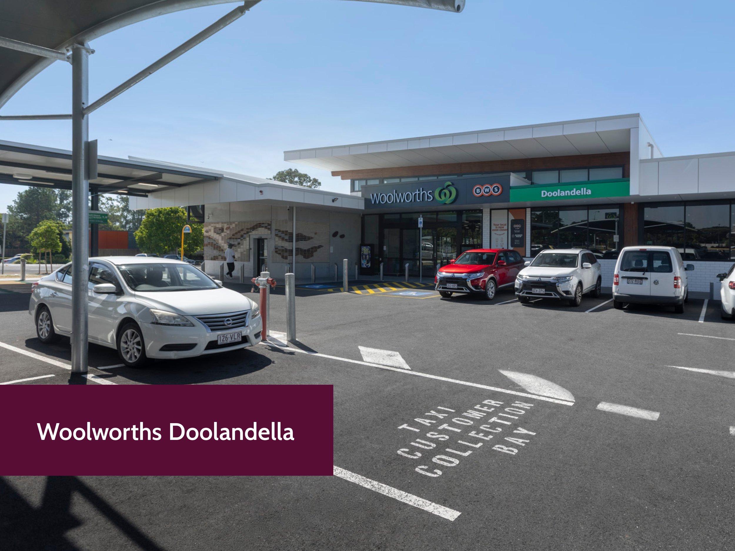 Exterior of Woolworths supermarket at Doolandella with parked cars in the parking lot, clear blue sky, and a person walking near the entrance.