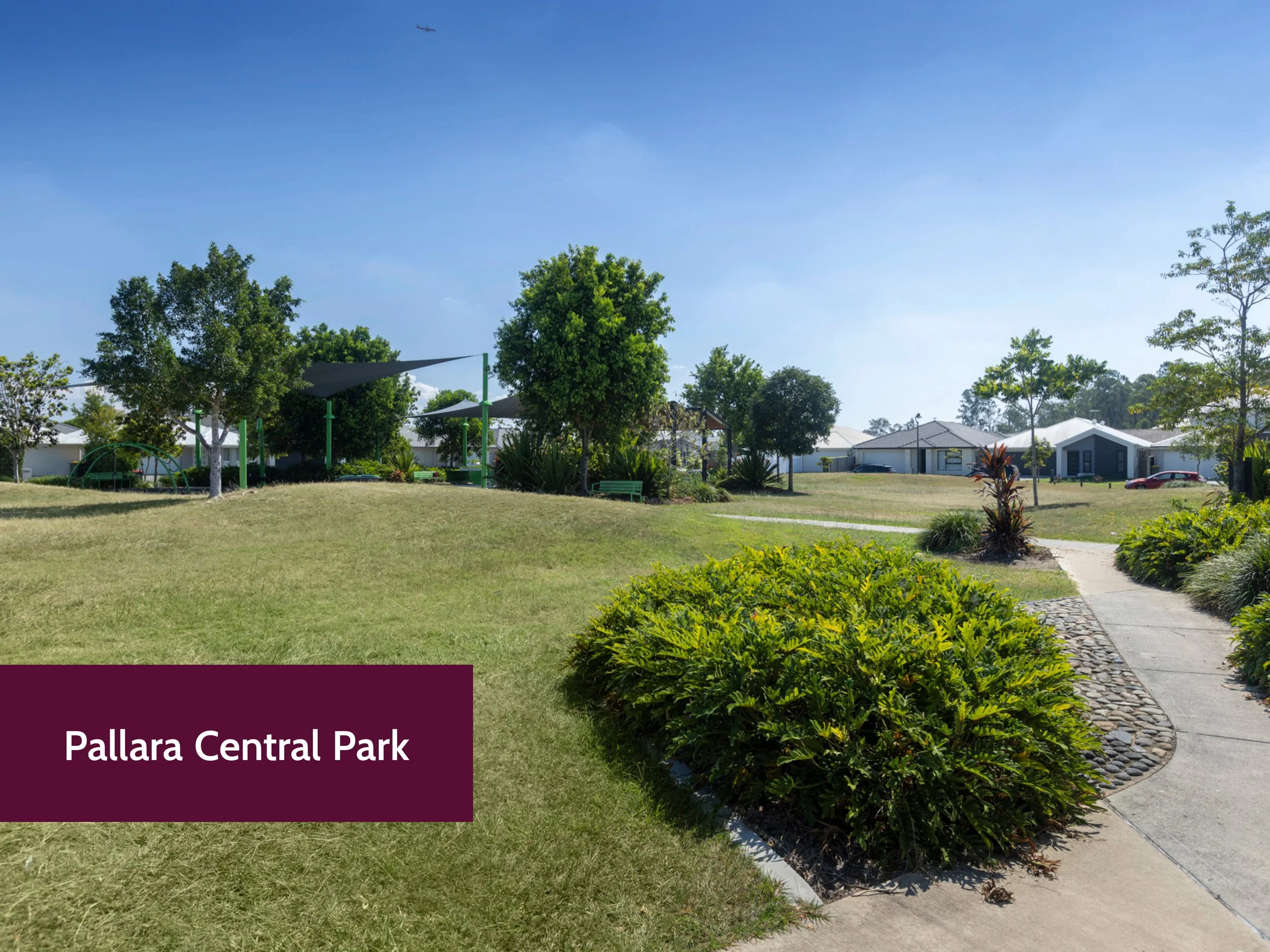 View of Pallara Central Park with lush green grass, trees, walking path, and a playground structure under a clear blue sky, with airplane visible in the distance.