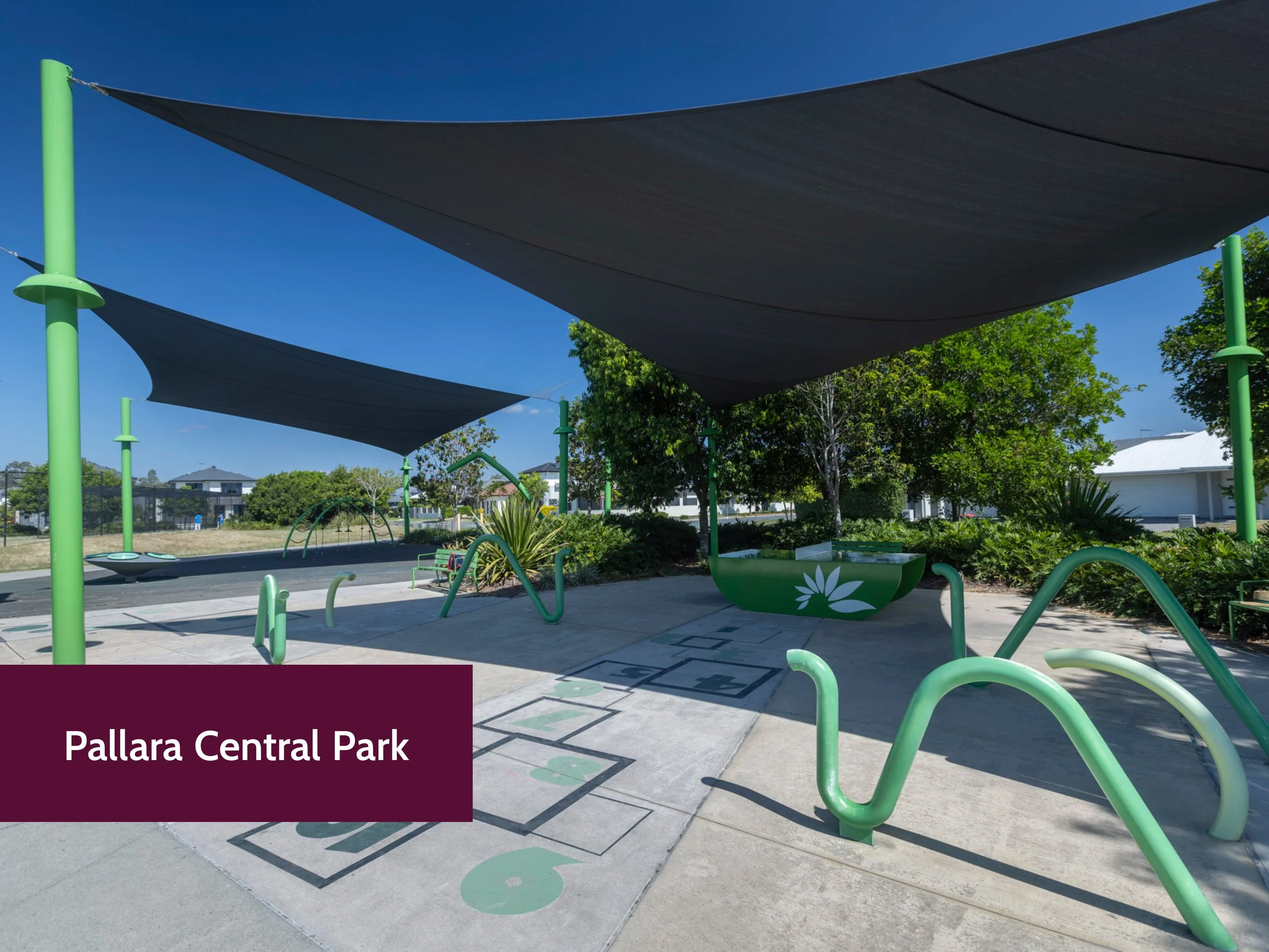 Shade sails over a playground area with green equipment, green poles, and a clear blue sky at Pallara Central Park, with trees and residential houses in the background.