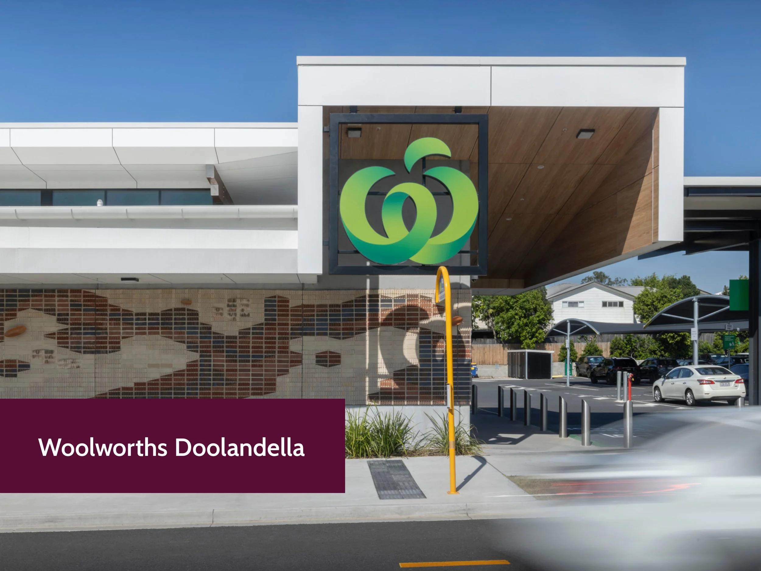 Exterior view of Woolworths Doolandella supermarket showing store signage, parking lot with several cars, and a landscaped area with trees.