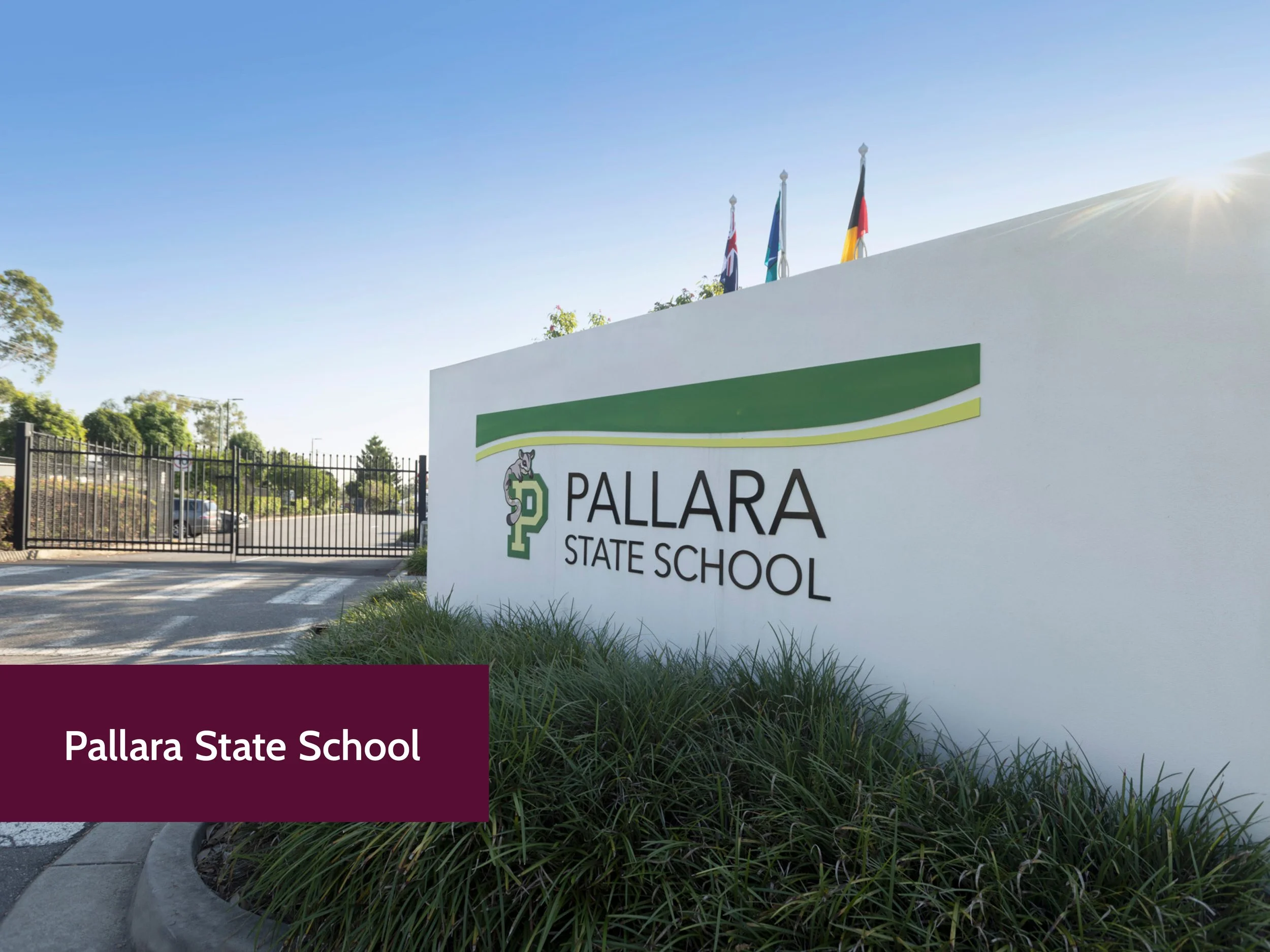 A white wall with a Pawlara State School sign, which includes a green, yellow, and black logo, and four flags on poles in the background against a blue sky with sunlight.