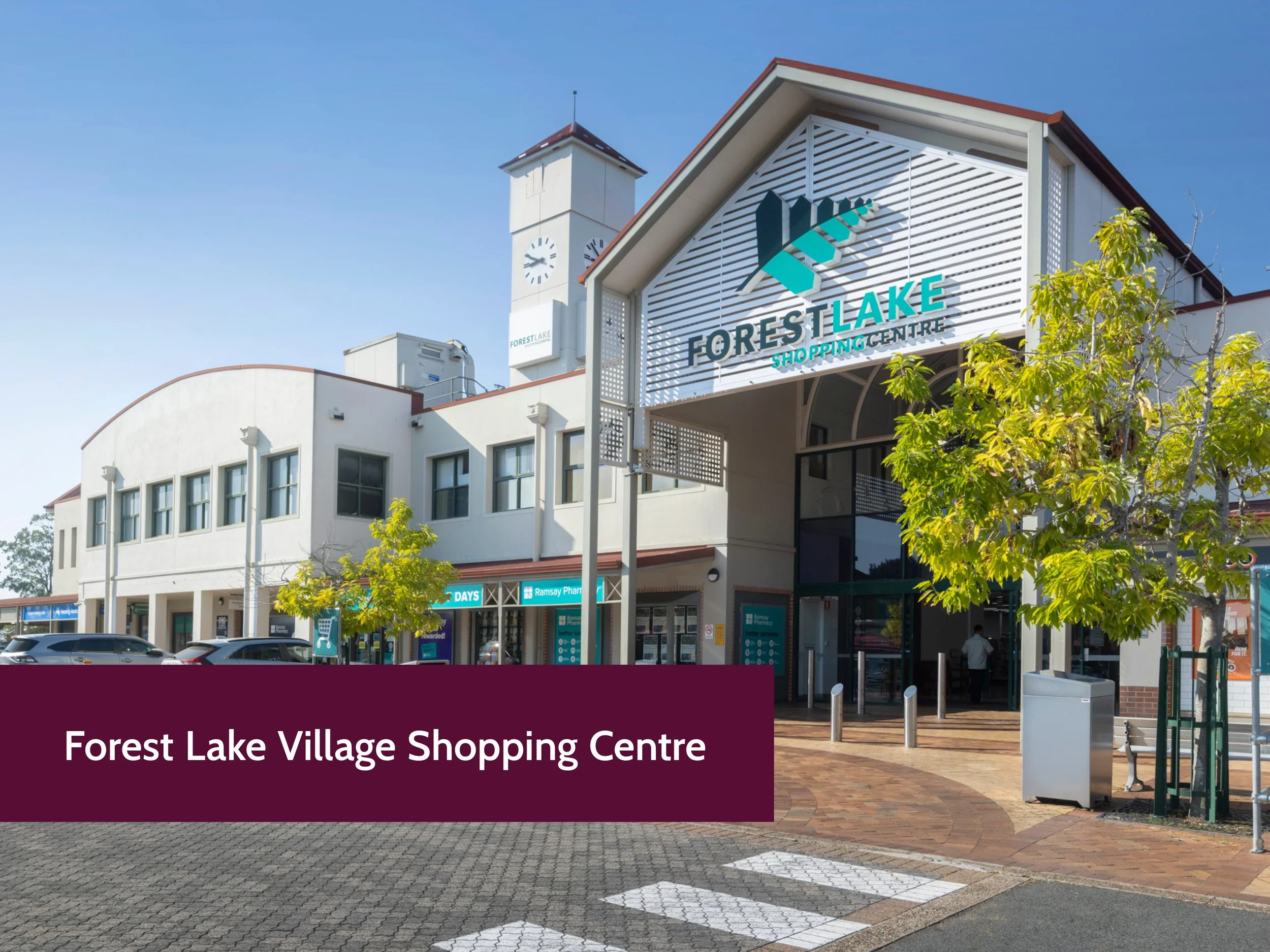 Exterior of the Forest Lake Village Shopping Centre with a large sign and a clock tower against a clear blue sky. There are trees, parked cars, and retail stores visible in the foreground.