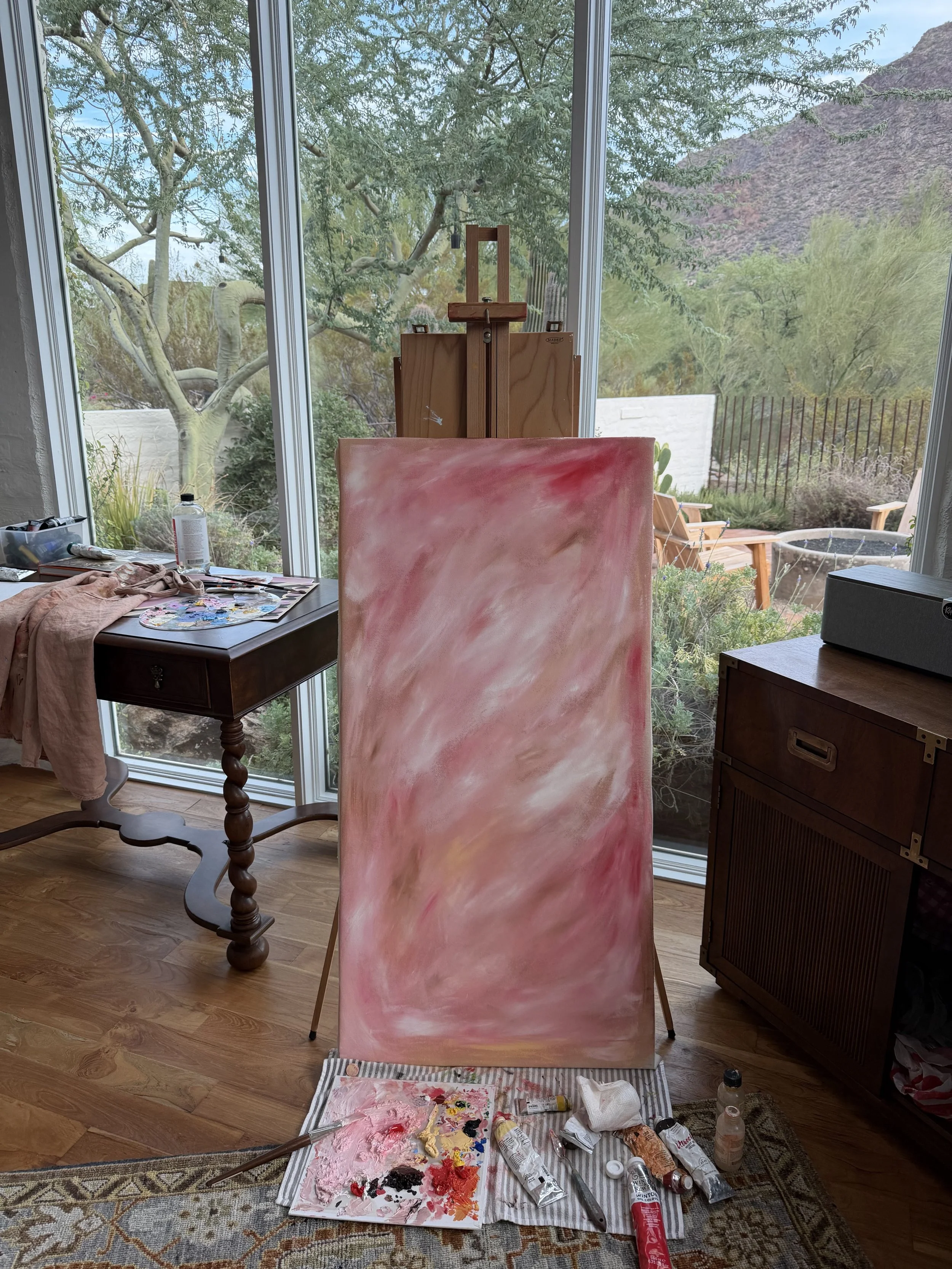 Sophia Grace Ashton's art studio with a pink abstract painting on a stand, a palette with various paint colors, and tubes of acrylic paint, brushes, and paper towels on a cloth-covered table in front of a large window overlooking trees and mountains.
