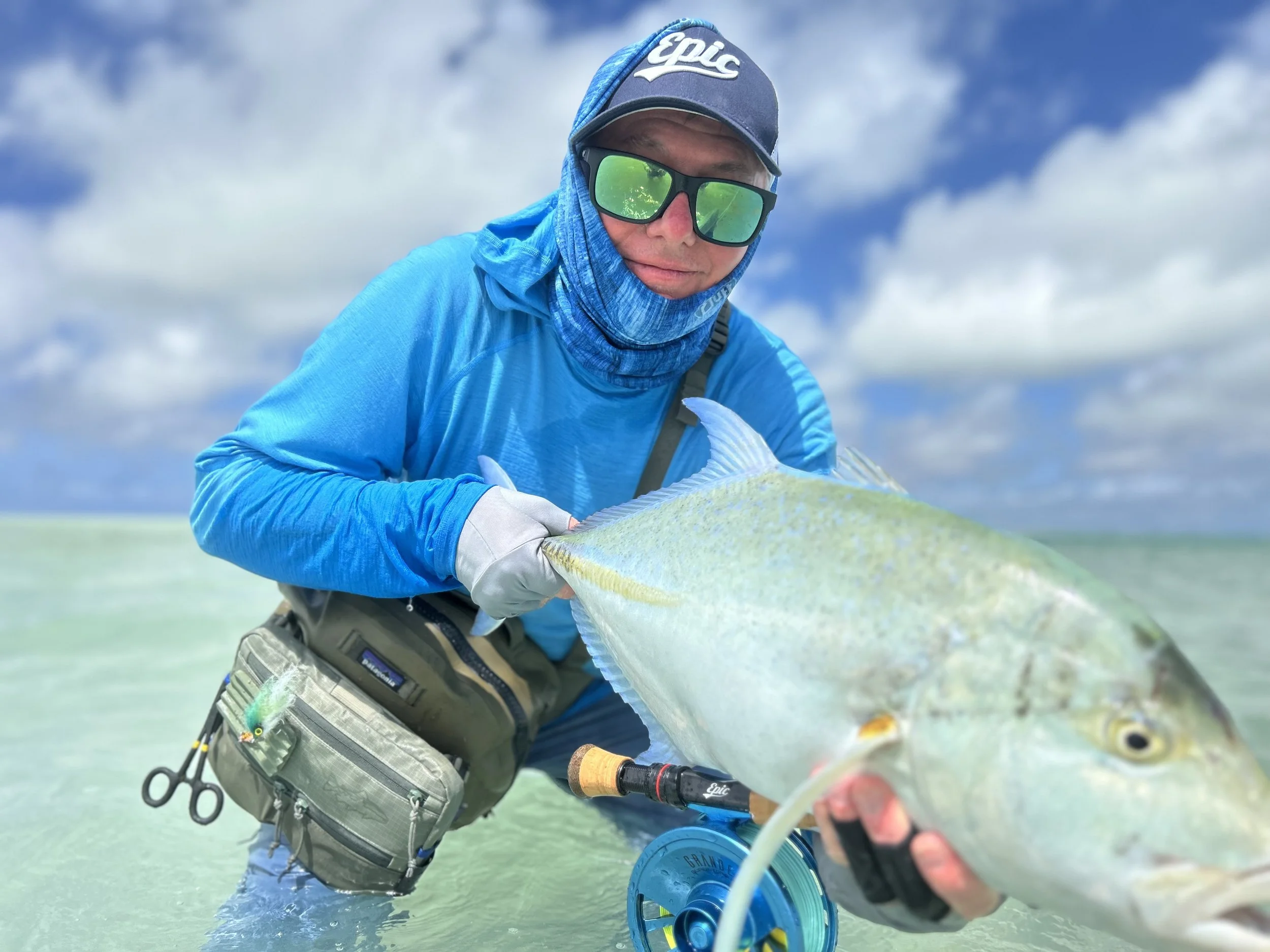 A person wearing a blue hoodie, sunglasses, and a branded fly fishing cap holding a large fish in the Kiritimati Lagoon under cloudy sky.