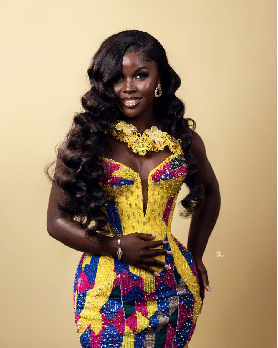 A woman with long, wavy hair in colorful traditional African attire, standing against a plain background.
