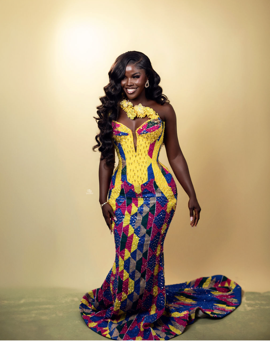 A woman with long curly black hair and dark skin wearing a colorful geometric-patterned gown with a train, standing against a plain yellow background.