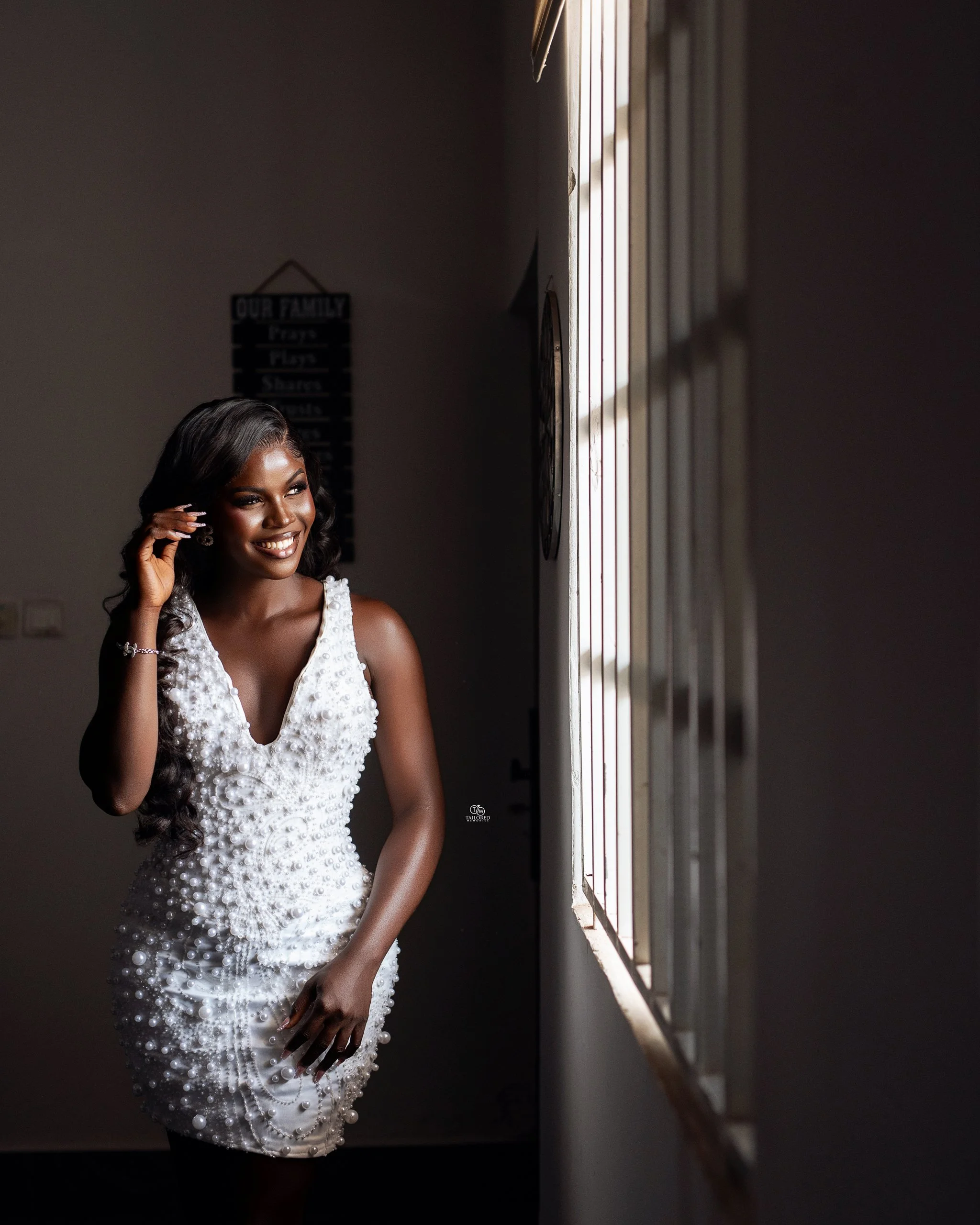A woman with dark skin and long black hair smiling, wearing a white dress decorated with pearls, standing next to a window with blinds, in a dimly lit room.