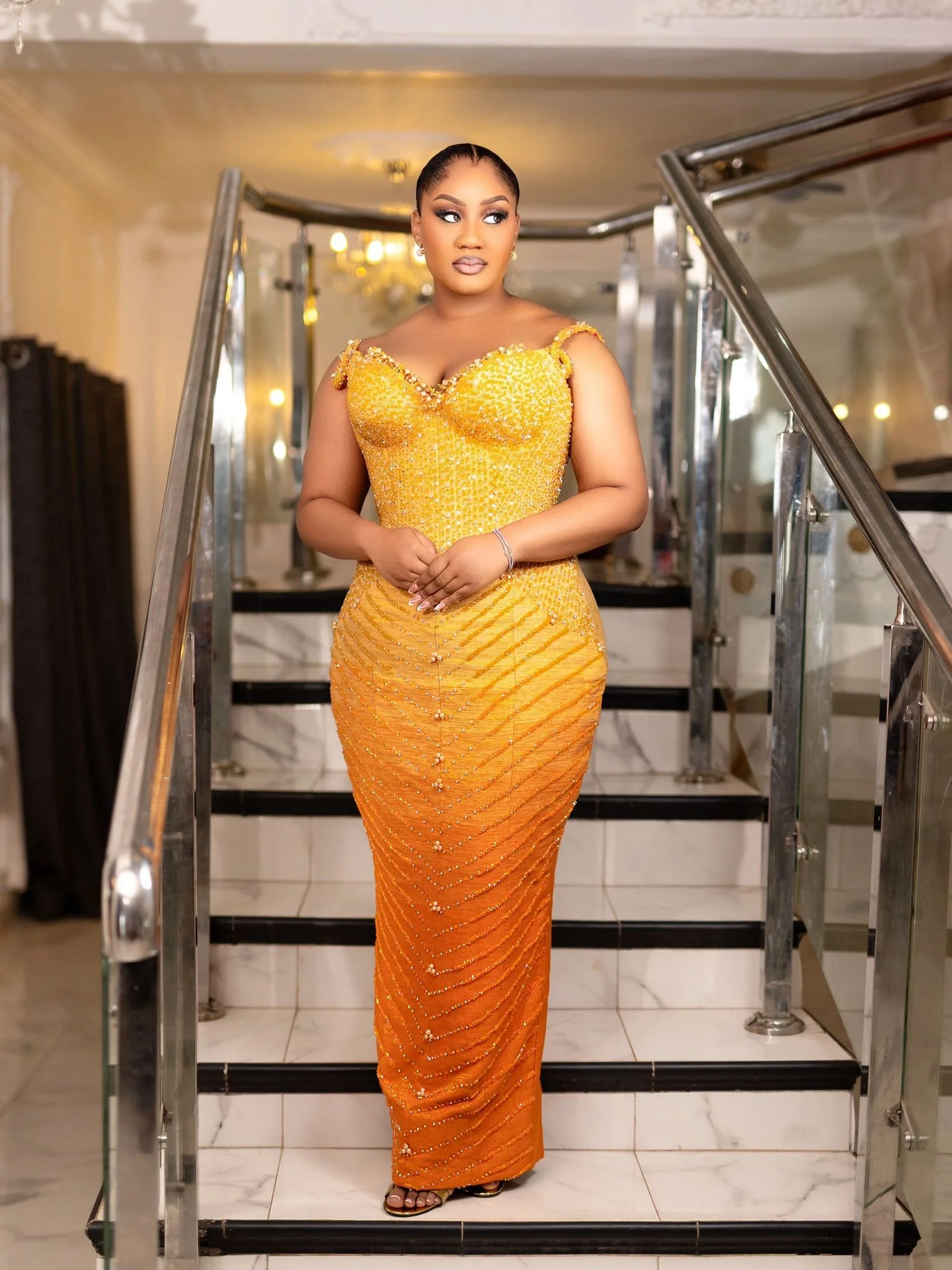 A woman in a shiny orange gown standing on a staircase with glass railings.