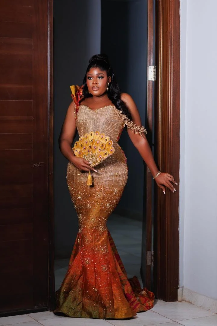 A woman in an elegant gold and orange sequined dress holding a decorated fan, standing in a doorway.