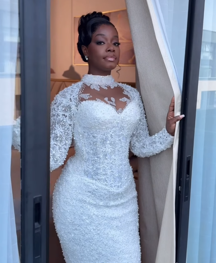 A woman in an elegant white wedding dress standing by a window and holding a curtain.