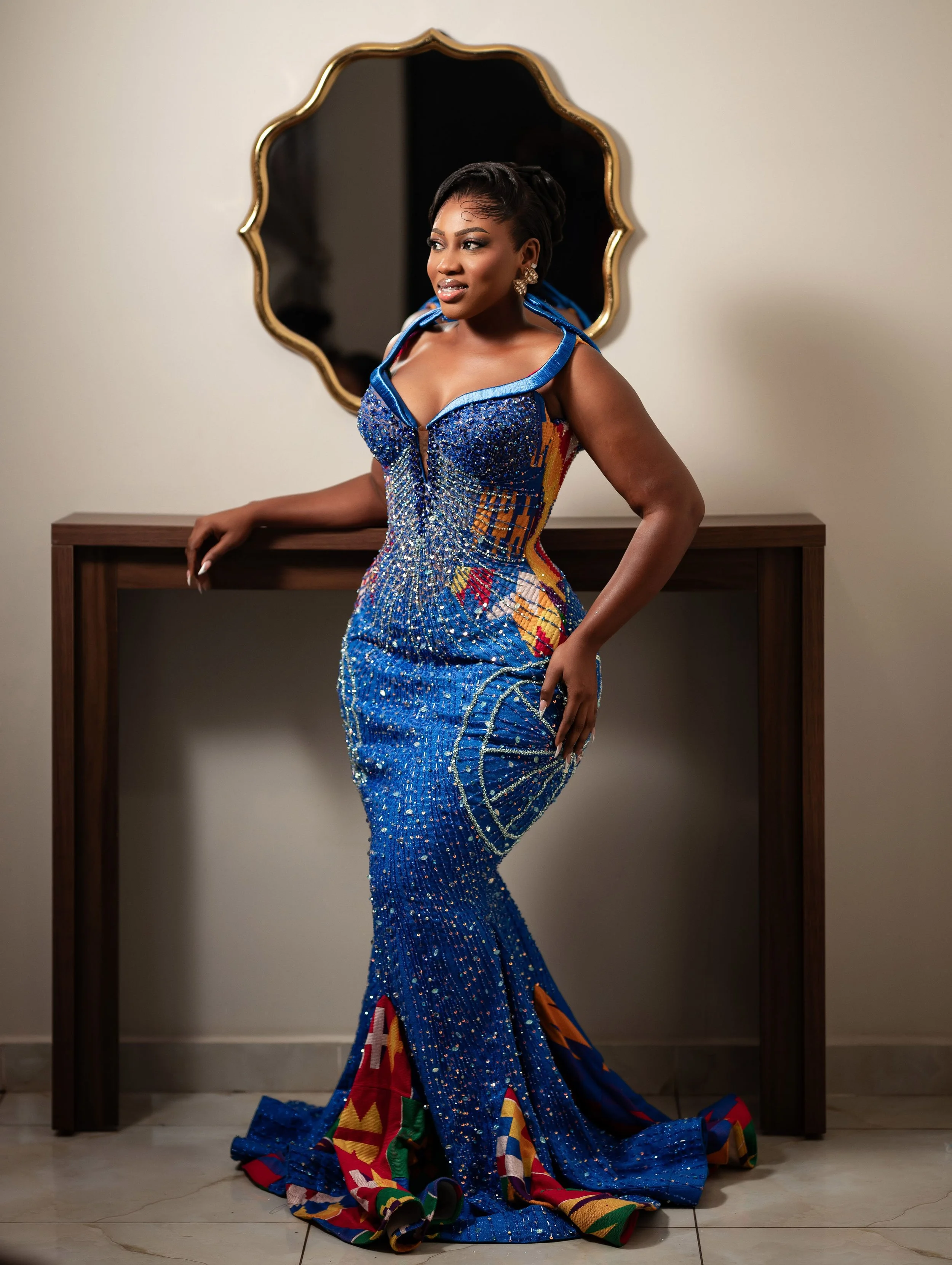 A woman wearing an elaborate, colorful, and sparkling blue dress with multicolored accents poses in front of a mirror and a plain wall.