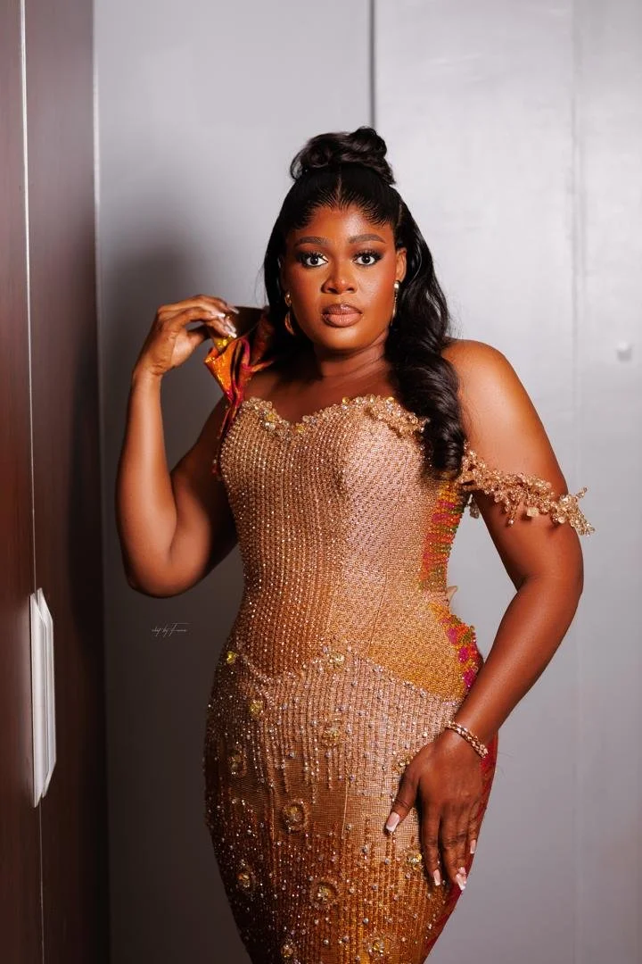 A woman with long black hair styled in an updo and loose waves, wearing a glamorous, fitted, gold-colored dress adorned with beads and sequins. She is standing against a plain wall, posing with one hand near her shoulder and the other resting on her 
