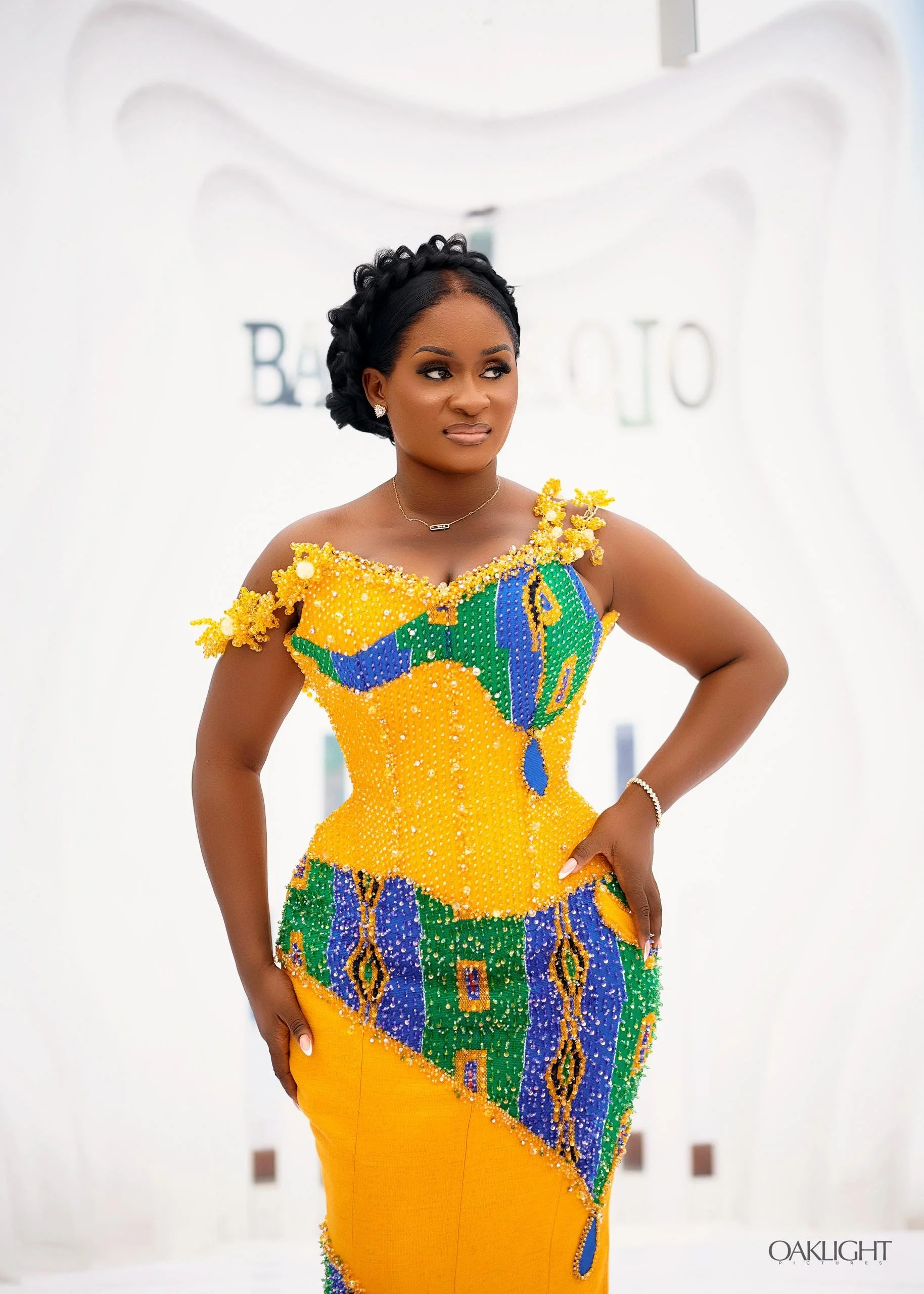A woman wearing a vibrant, colorful dress made of beads and fabric, with her right hand on her hip. She has dark skin, styled black hair, and wears earrings, a necklace, and a bracelet. The background is white with some blurred signage.