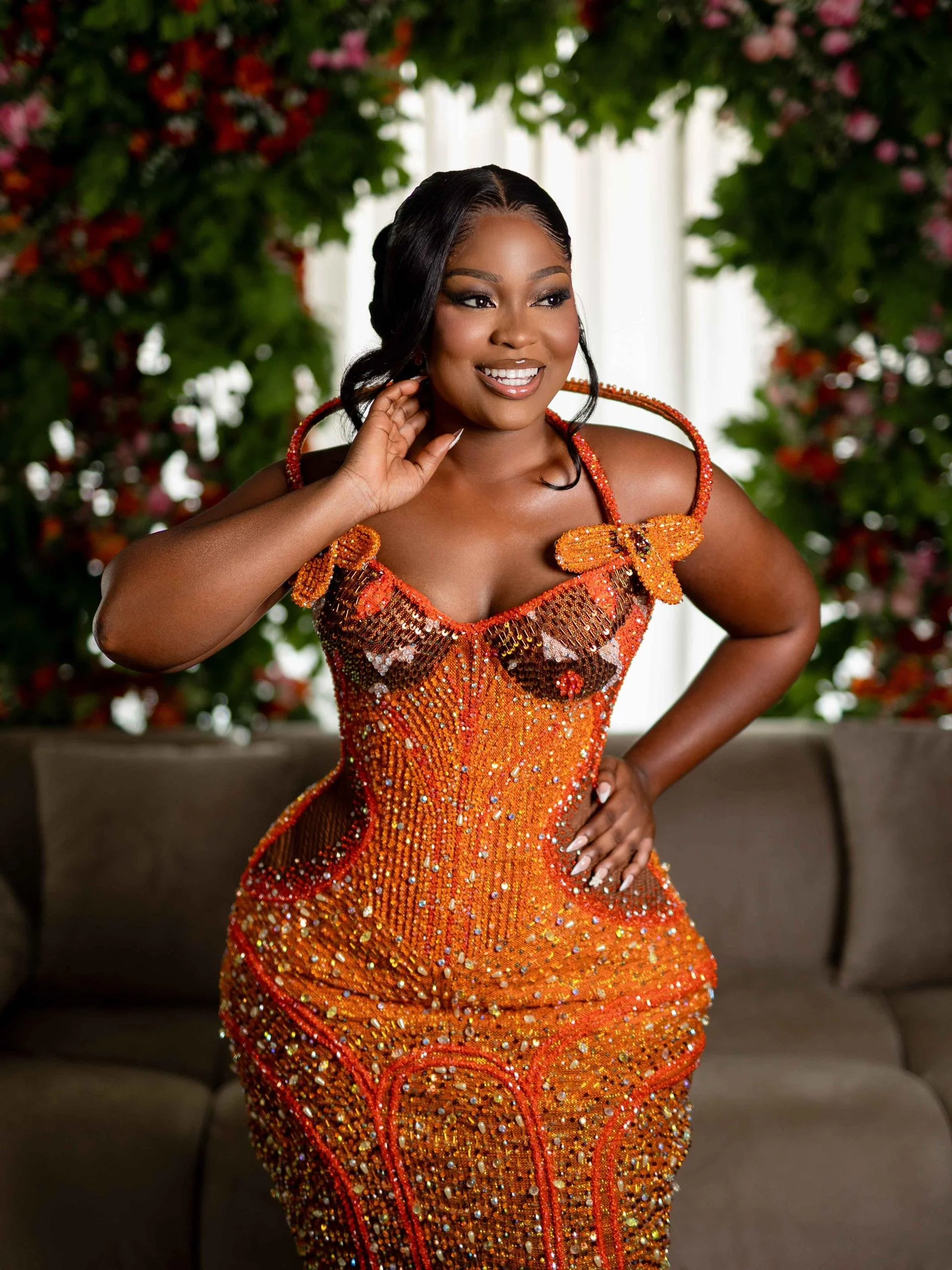A woman in a vibrant orange, sequined dress with butterfly-shaped shoulder decorations, smiling, with her hand near her face, in a setting with greenery and pink flowers in the background.