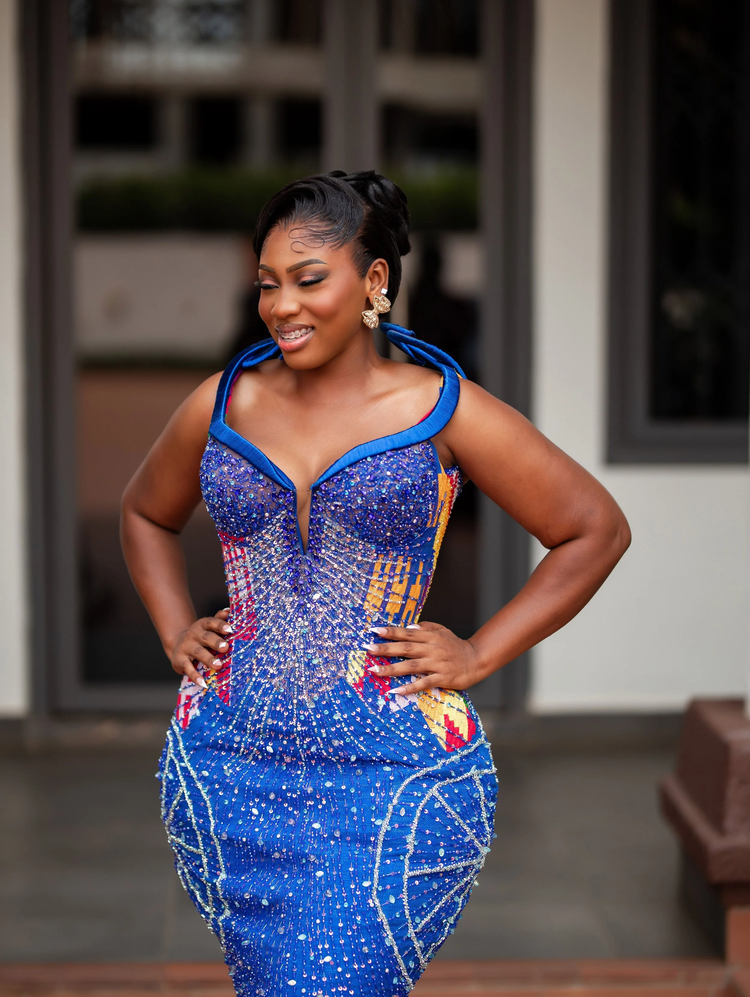 Woman dressed in a vibrant, intricately beaded blue and multicolored gown, smiling with her eyes closed, standing outdoors.