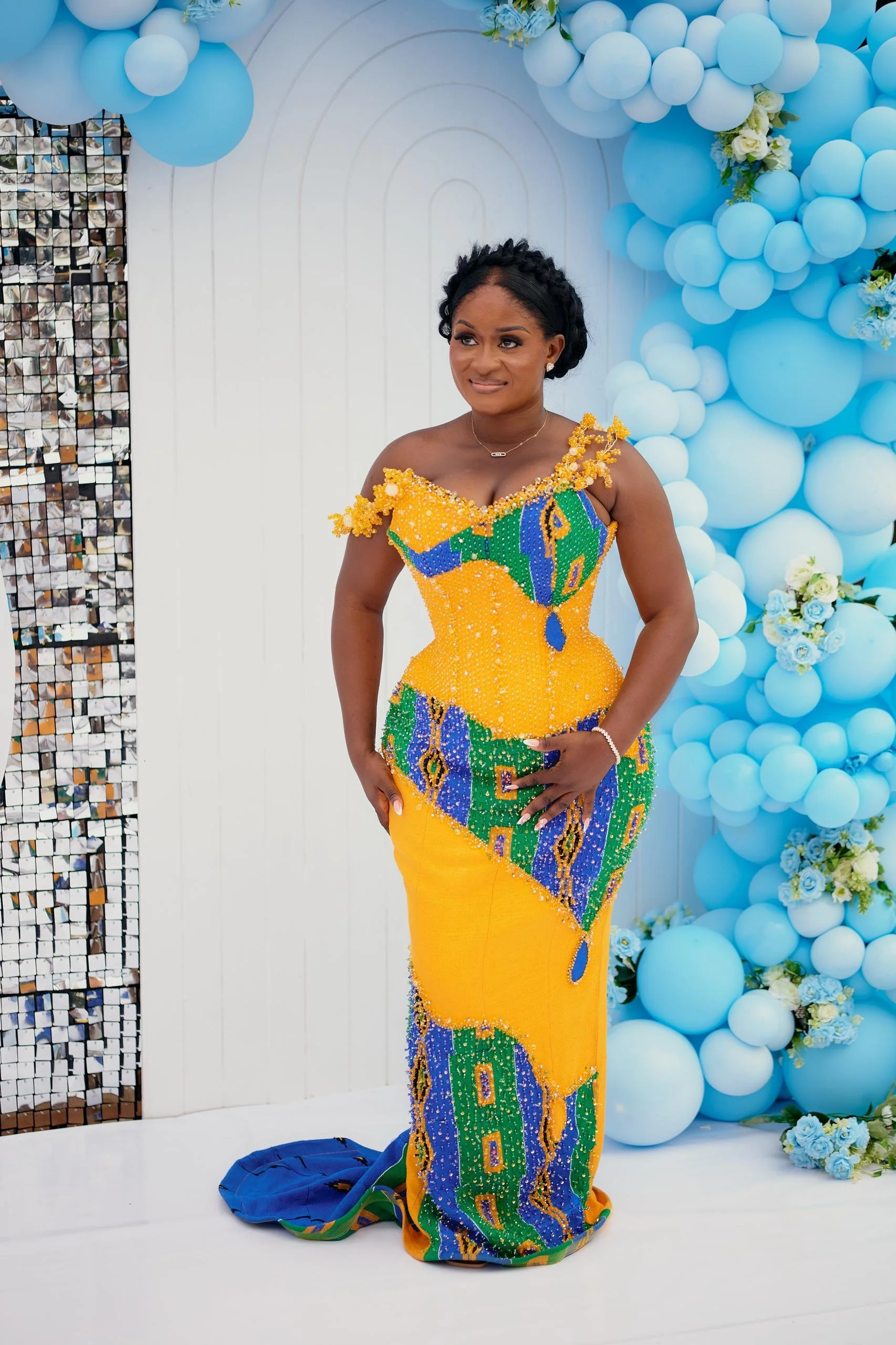 A woman in a brightly colored traditional African dress, standing in front of a decorated backdrop with blue balloons and white flowers.