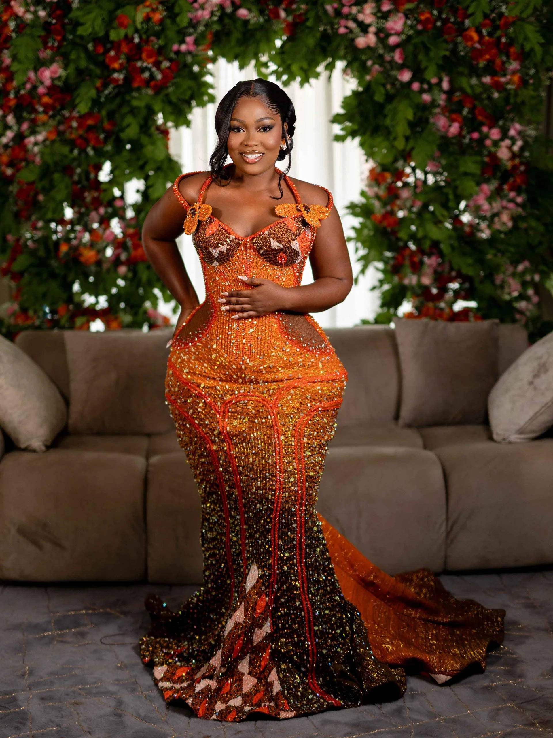 A woman in an elaborate, sparkling orange and gold gown with floral details is standing in front of a floral backdrop, smiling at the camera.