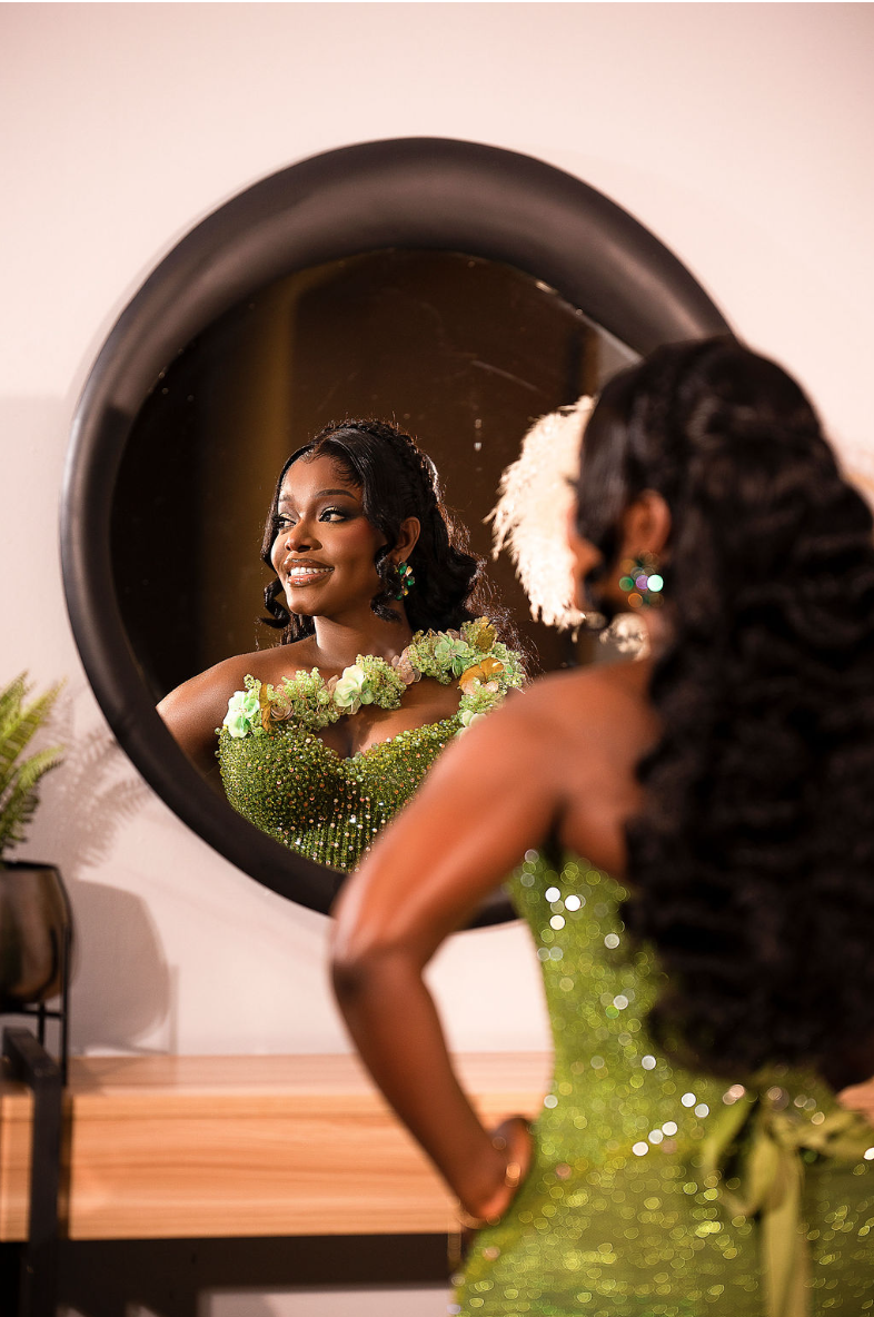 A woman in a green, beaded dress looks at her reflection in a mirror, smiling. She has earrings, and her hair is styled in waves with a white feathered accessory. The setting appears to be a dressing room with a wooden surface and a plant.
