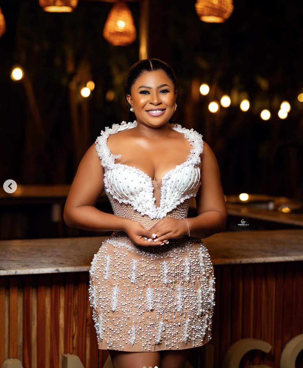 A woman in an ornate, embellished dress smiling at the camera in an outdoor setting with warm lighting.