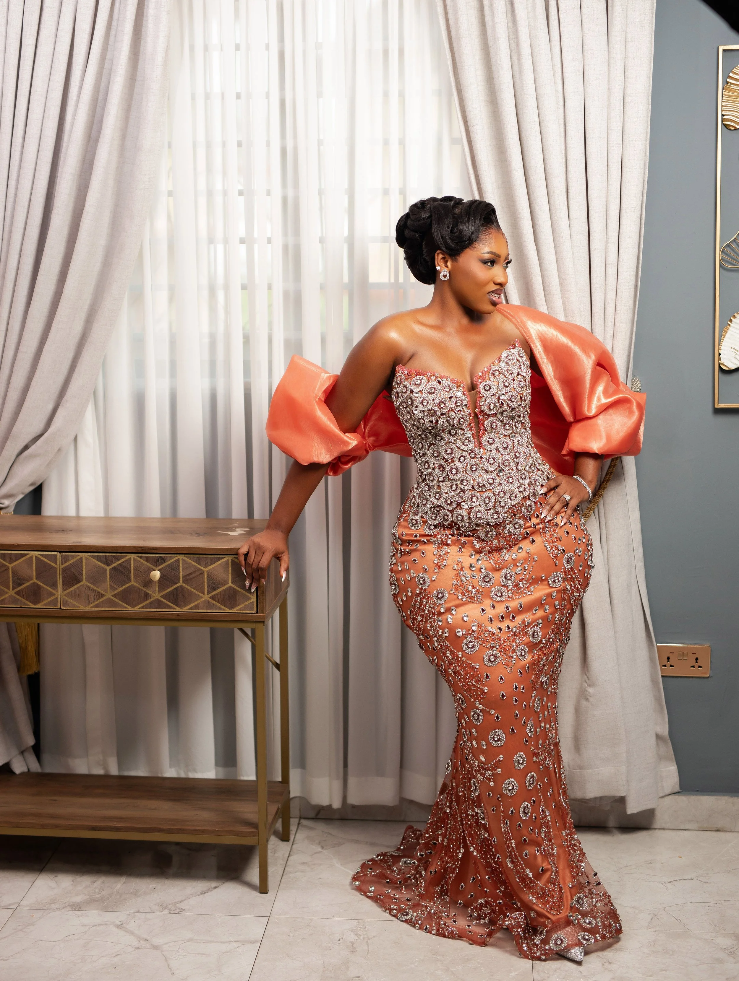 A woman dressed in an ornate, peach-colored gown is standing indoors near a window with light-colored curtains. She is posing with one hand resting on a wooden table and the other on her hip, looking to her left. The gown features intricate beadwork 