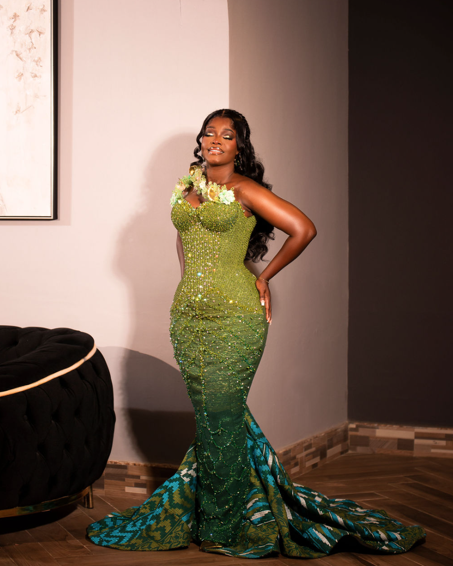 A woman in a fitted, glamorous green gown with embellishments and a lace-up train, standing indoors with one hand on her hip and a confident expression.