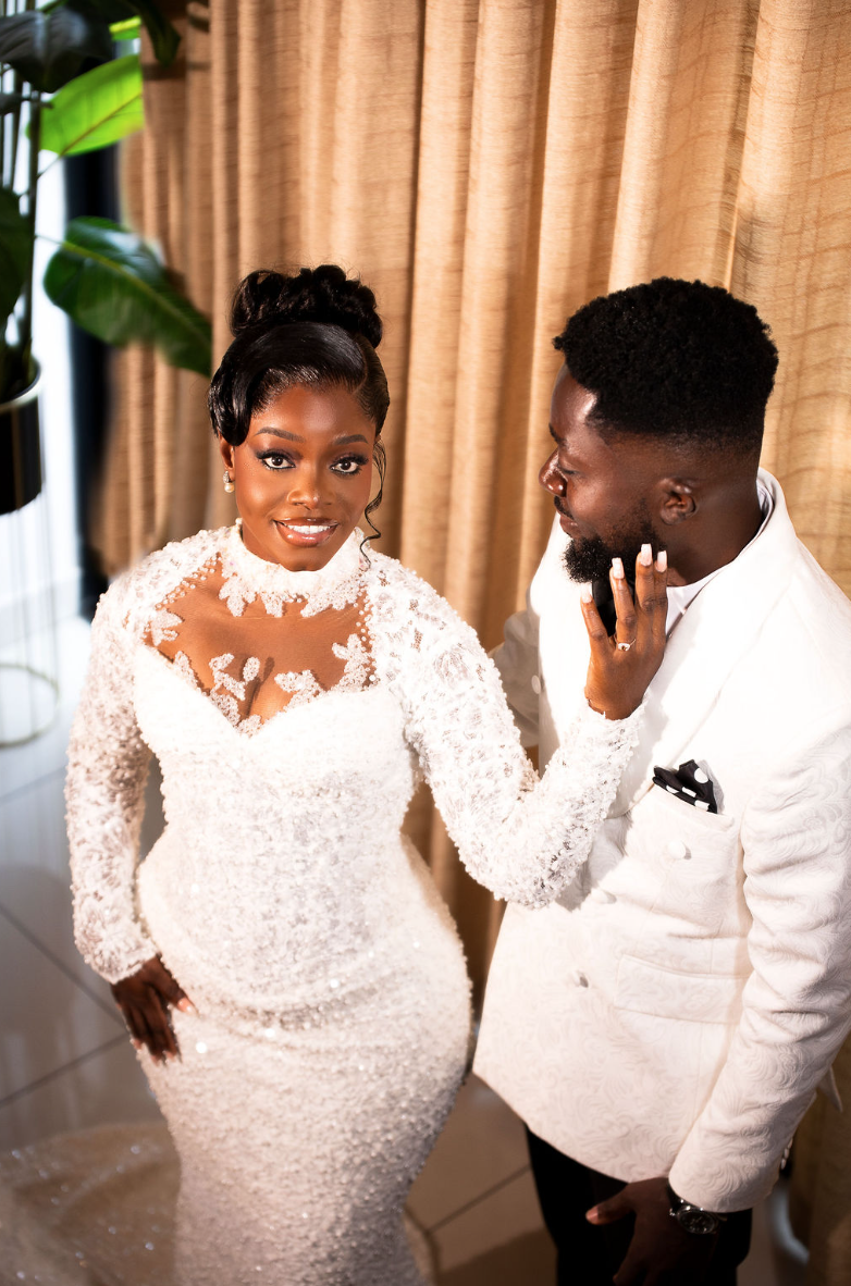 Bride in a white lace wedding dress smiling at the camera, groom in a white tuxedo with a black pocket square looking at her, in a room with beige curtains and a potted plant.