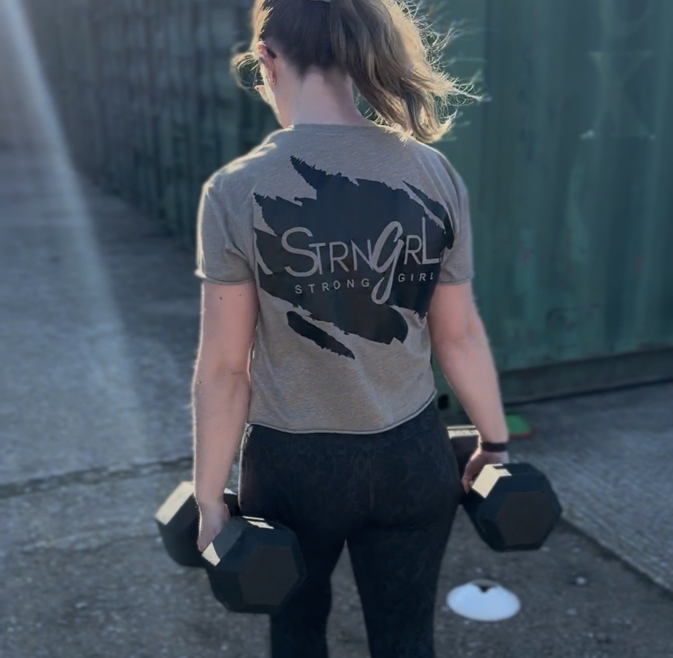 A woman in workout clothes holding two black dumbbells outdoors, wearing Strngrl Merchandise