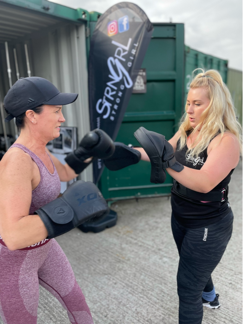 Two women practicing boxing outdoors, one in a purple sports outfit with boxing gloves, and the other in black athletic wear with boxing pads, near a banner for Strong Girl.