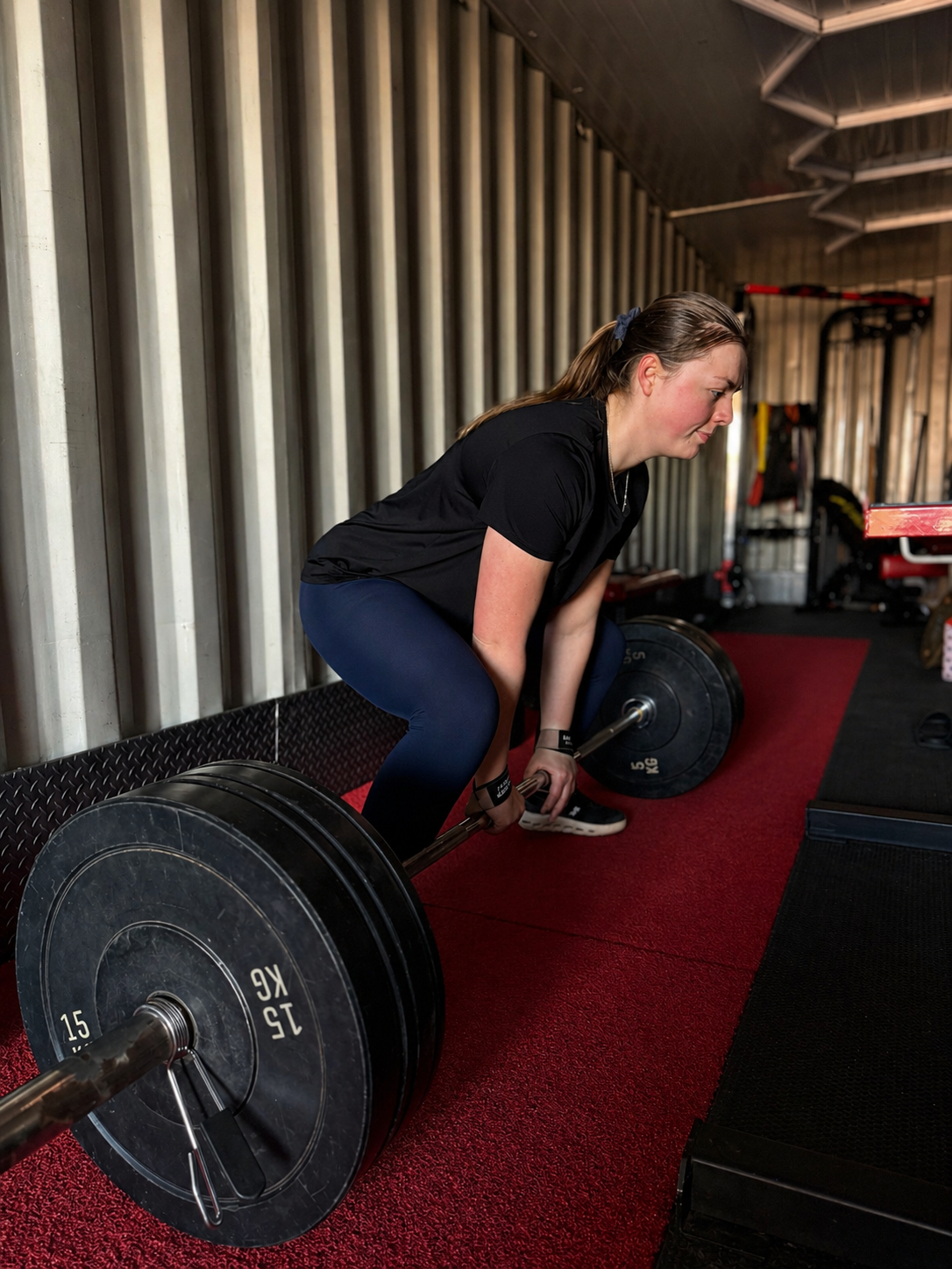 A woman performing deadlifts with a barbell in a gym, positioned on a red mat near a metal wall.
