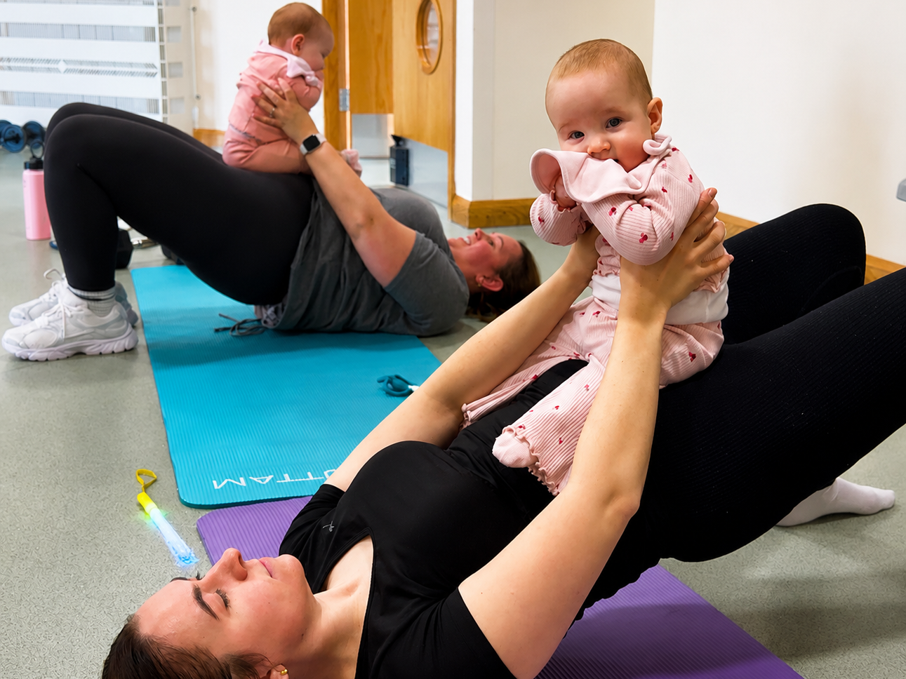 Two women exercising on yoga mats with babies, one on her back and one lying on her back holding a baby, in a fitness or exercise room.