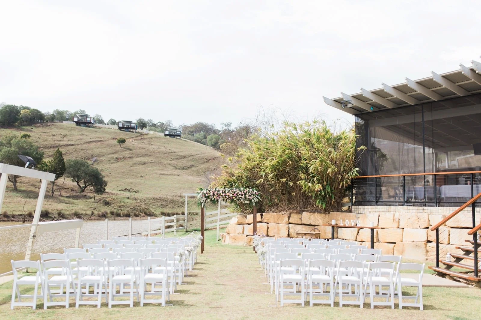 Outdoor wedding ceremony setup with white chairs arranged in rows on grass, floral arch at the altar, hillside with small buildings in the background, and a modern glass building on the right.